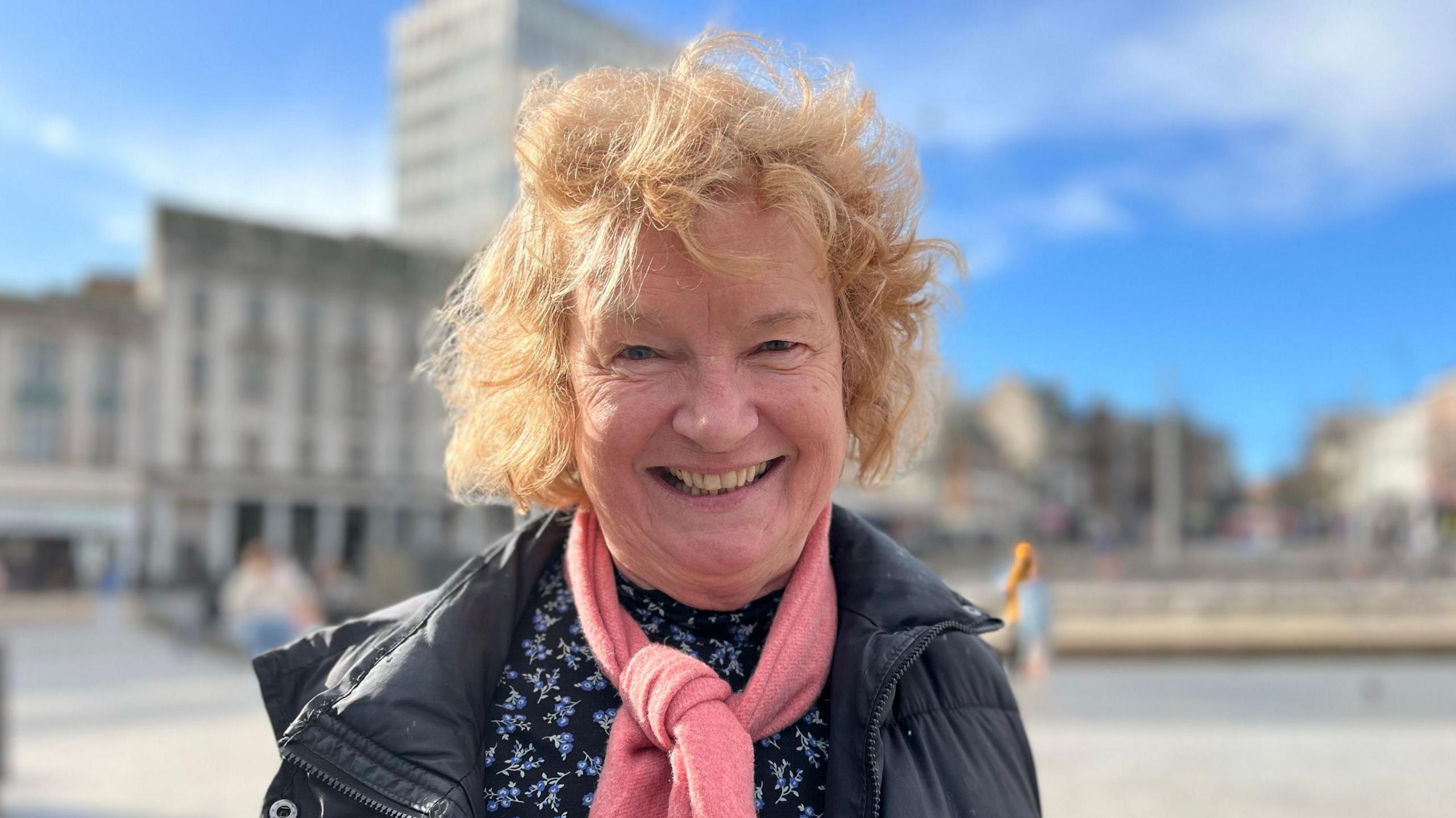 A white woman with dark blonde, short, curly hair, wearing a pink scarf and blue padded coat. She is smiling. 