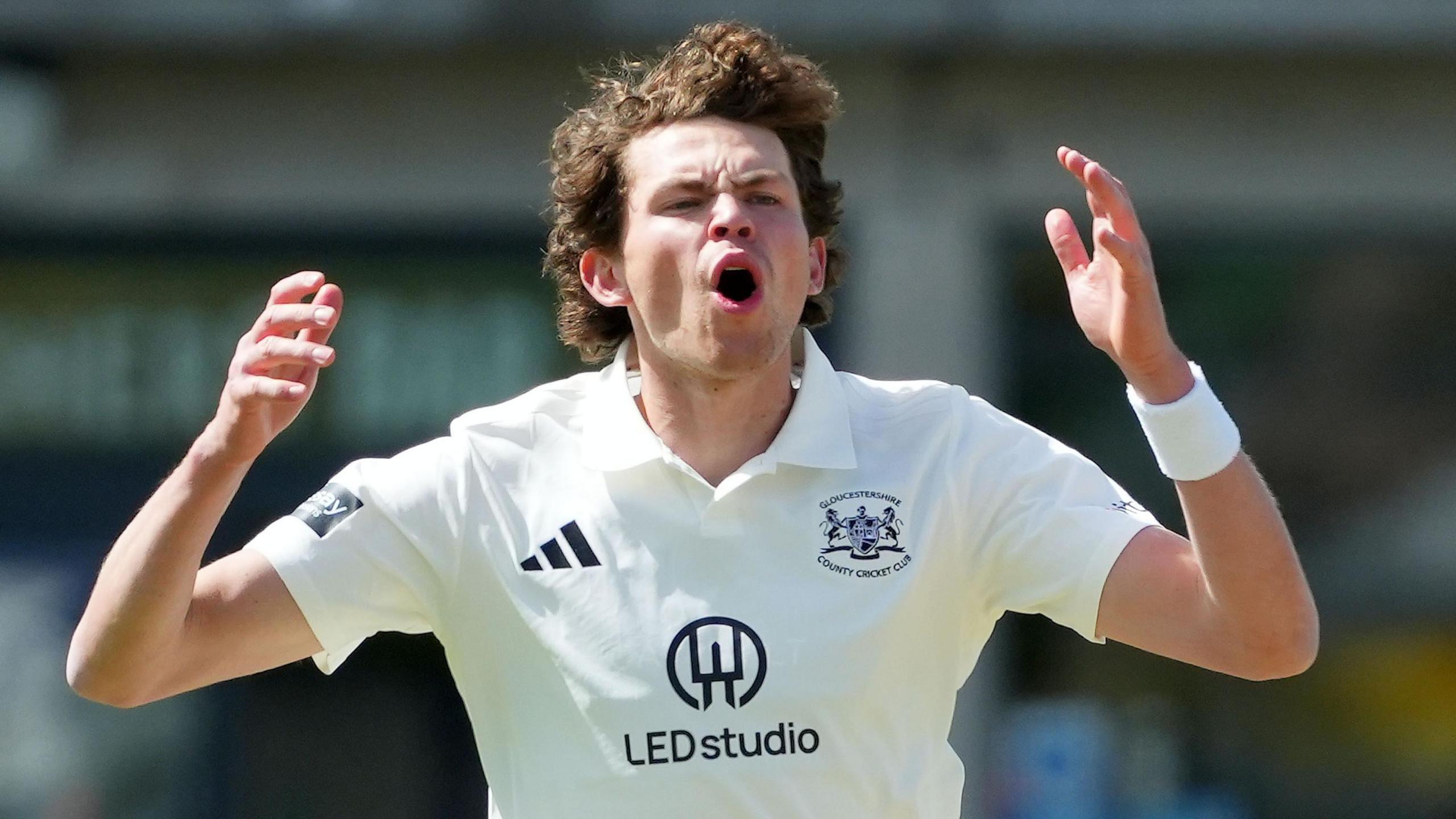 Henry Brookes, wearing a white Adidas cricket shirt with the Gloucestershire county logo, opens his mouth and puts his hands up by the side of his head after bowling a ball in the County Championship Division Two game against Lancashire