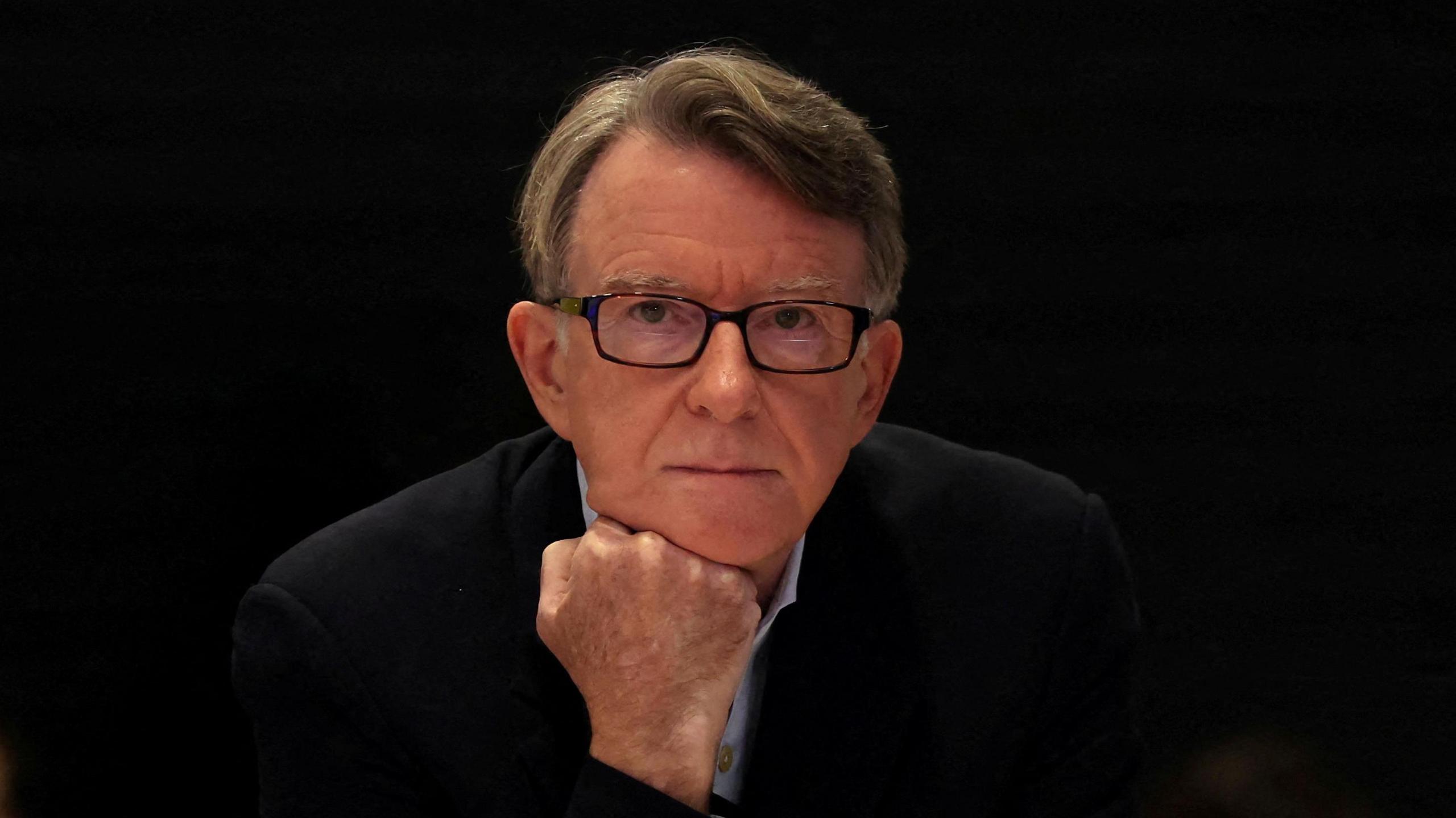Peter Mandelson is resting his chin on his fist. He has short brown hair and black square-framed glasses.