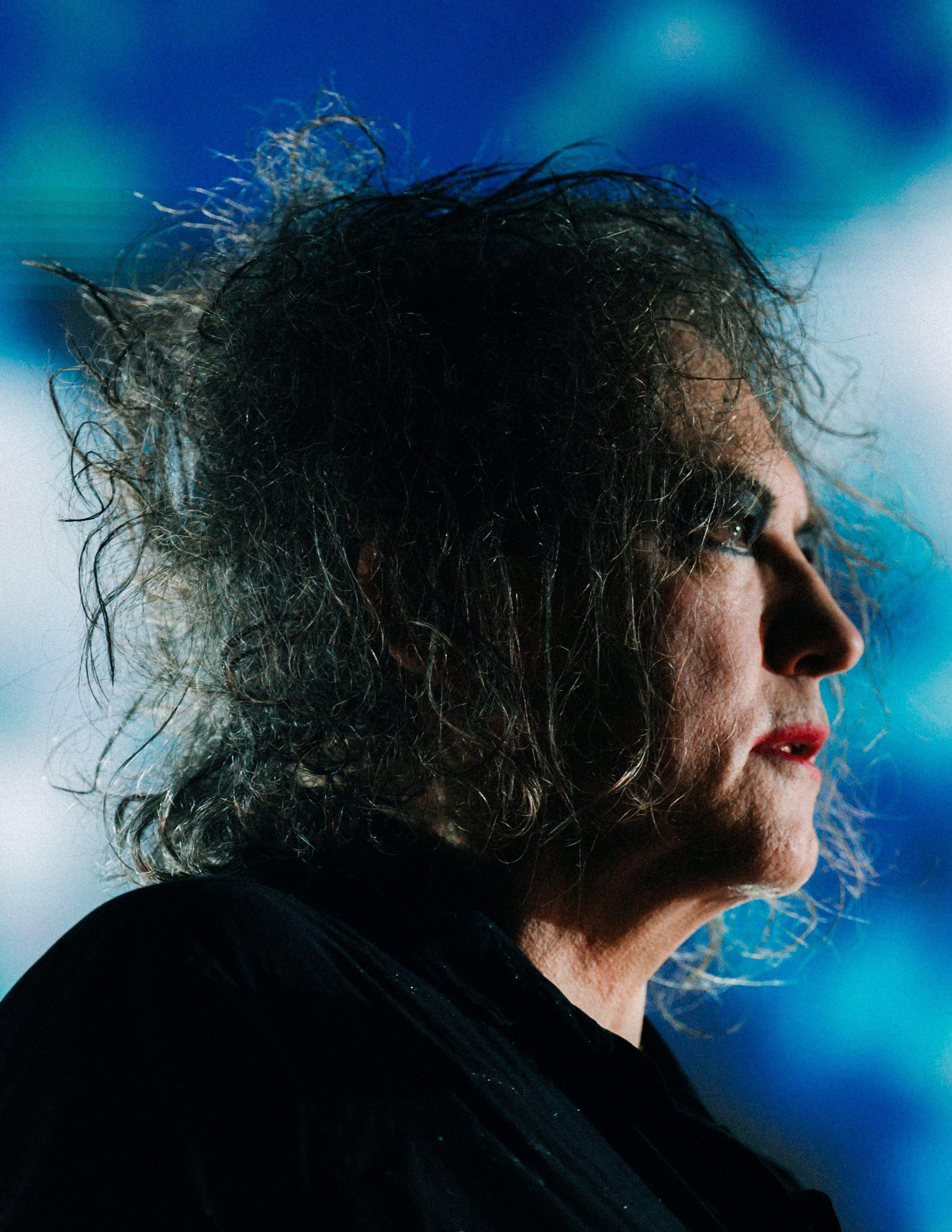 Profile shot of The Cure's Robert Smith against a blue background. Smith is wearing black eye make up and red lipstick.