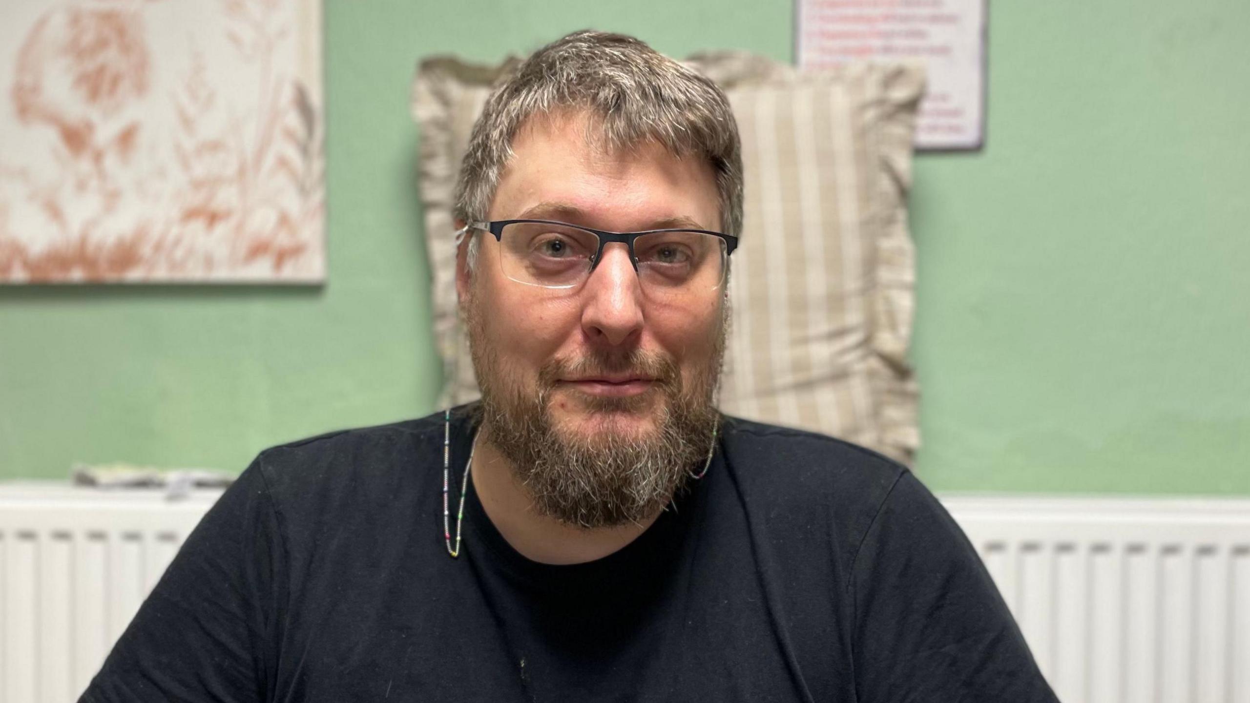 Head and shoulders image of Jason Bibby, with a blue shirt and glasses. In the background is a white radiator and green wall.