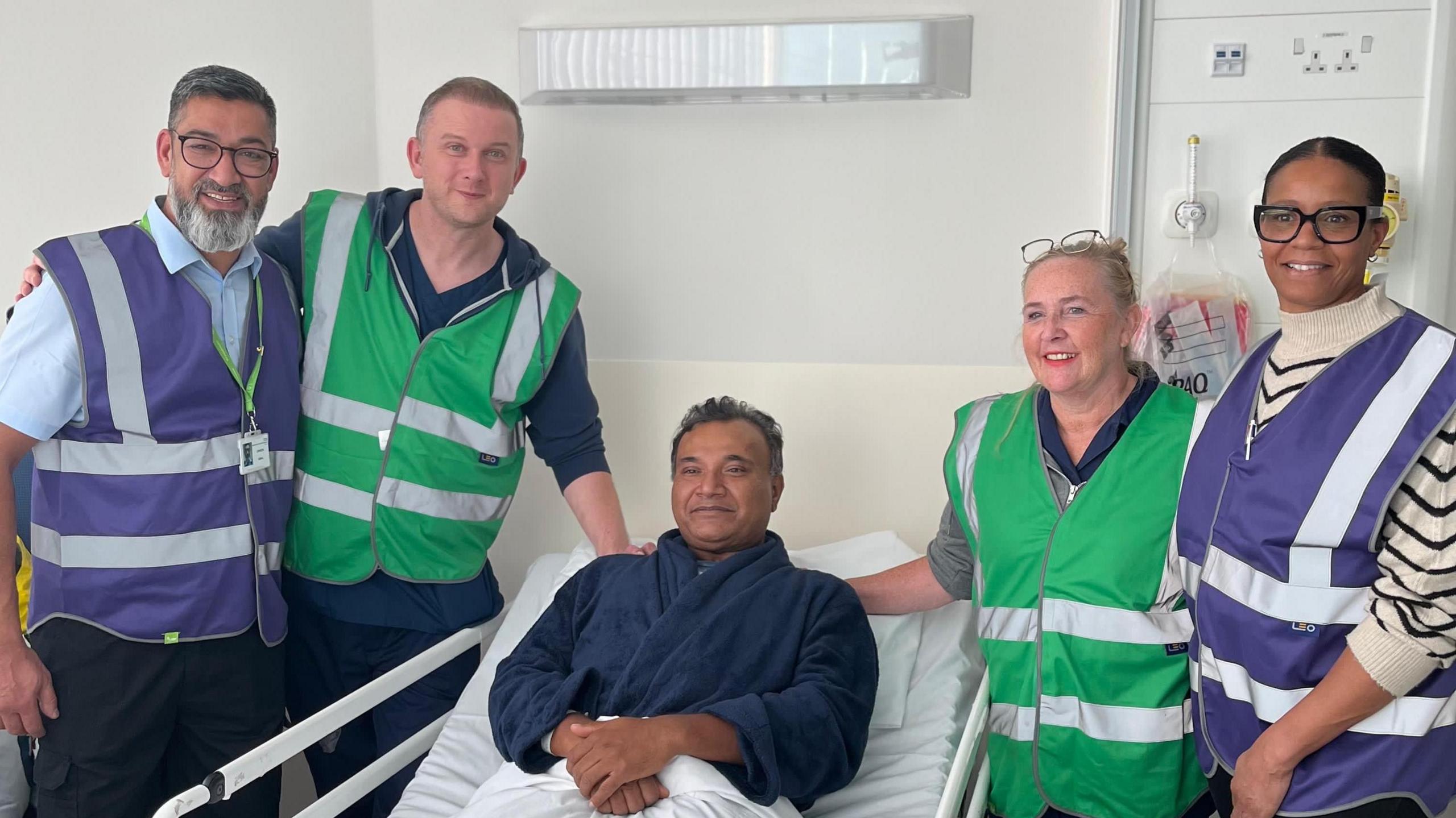 A man sits upright in a hospital bed, with two men in hi-viz tops standing to the left of the bed, and two women in hi-viz tops standing to the right 