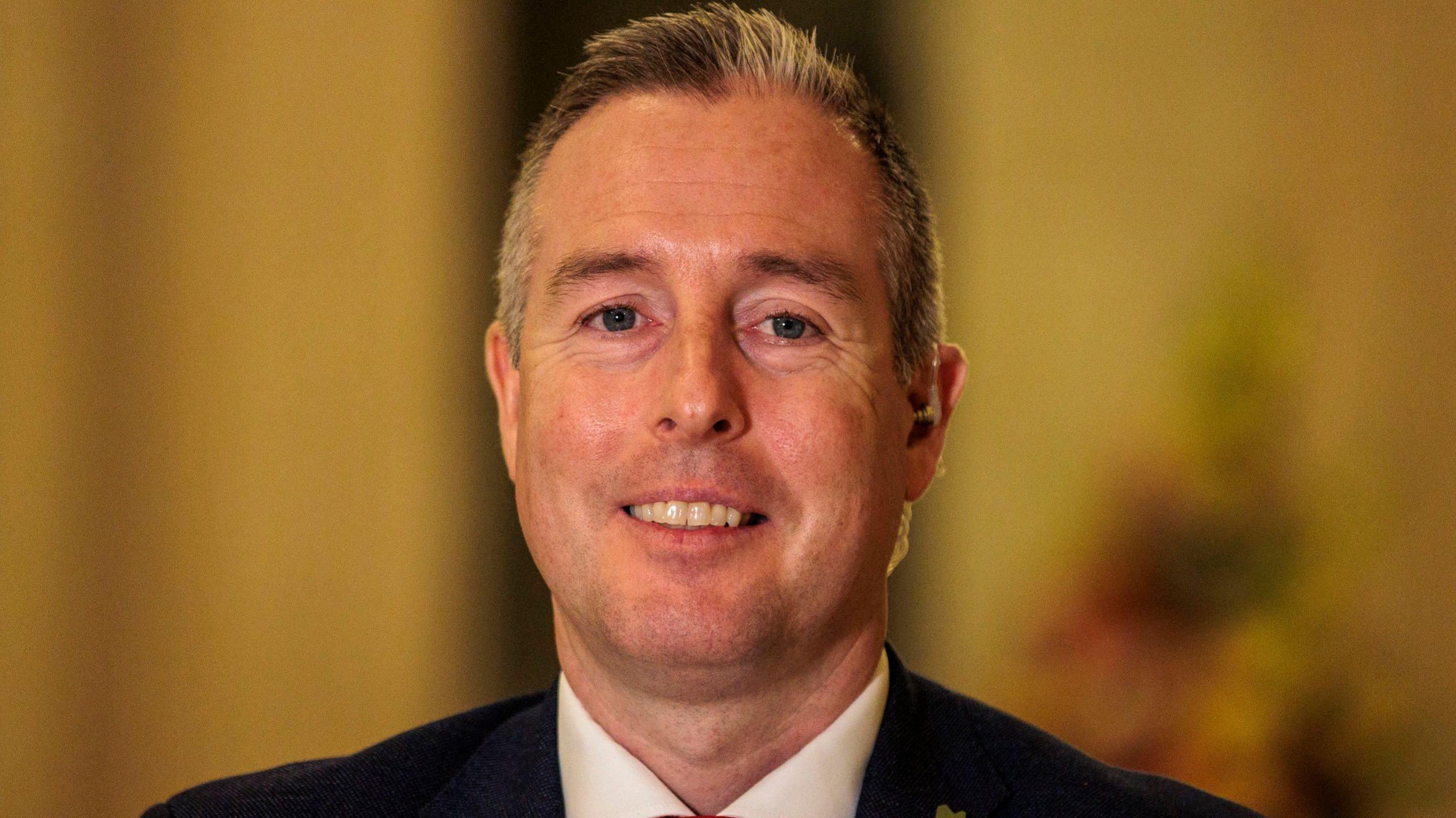 Paul Givan smiling at the camera, he has greyish black hair, he is wearing a black blazer and white shirt