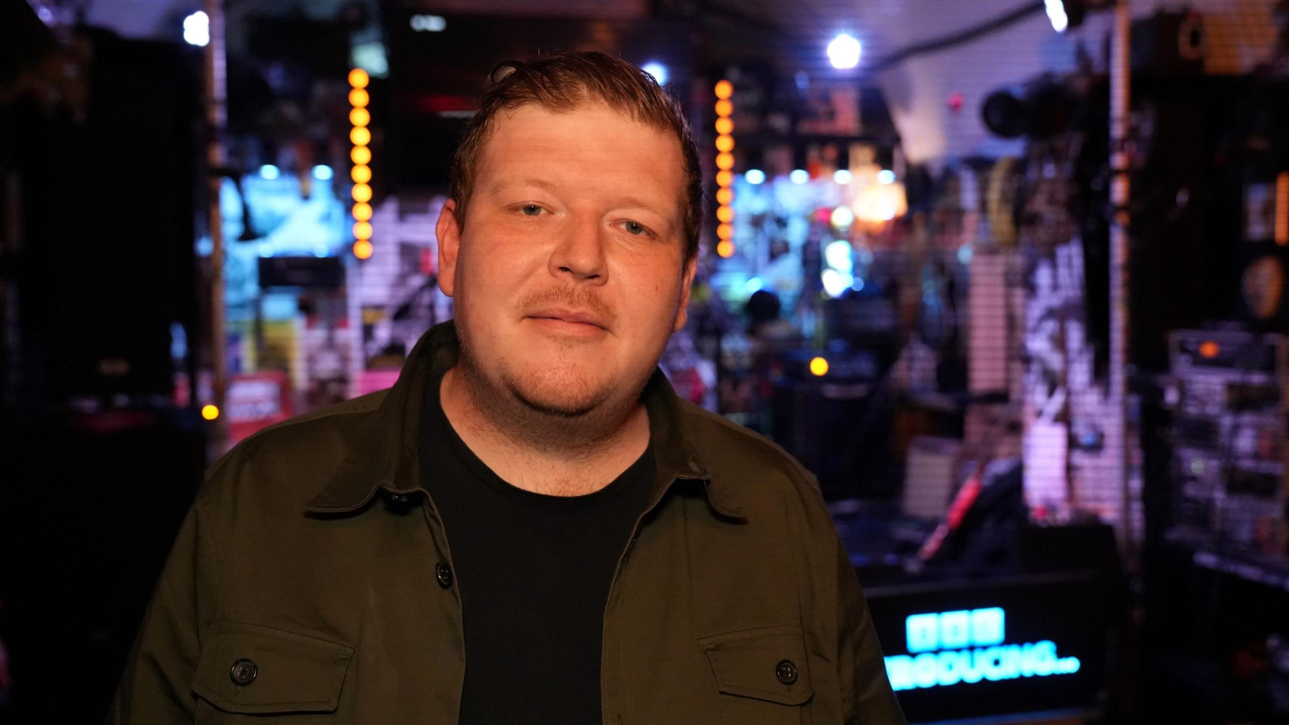 A man smiling at the camera. He is wearing a black top with a dark green shirt over the top. There is a stage behind him.