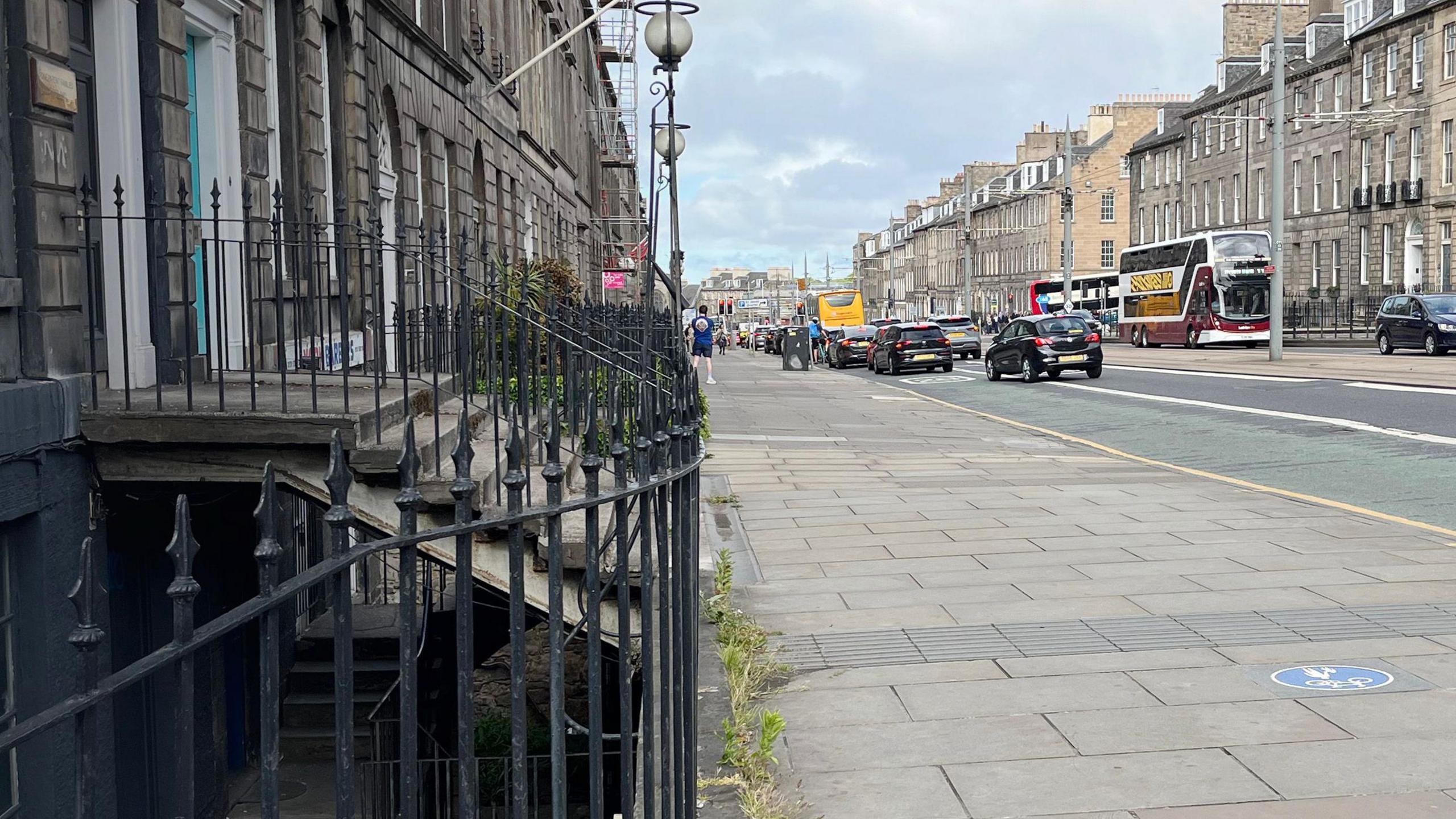 A street with buses and cars on the road, a wide stone slab pavement, and railings and an area in shadown below steps to the left.