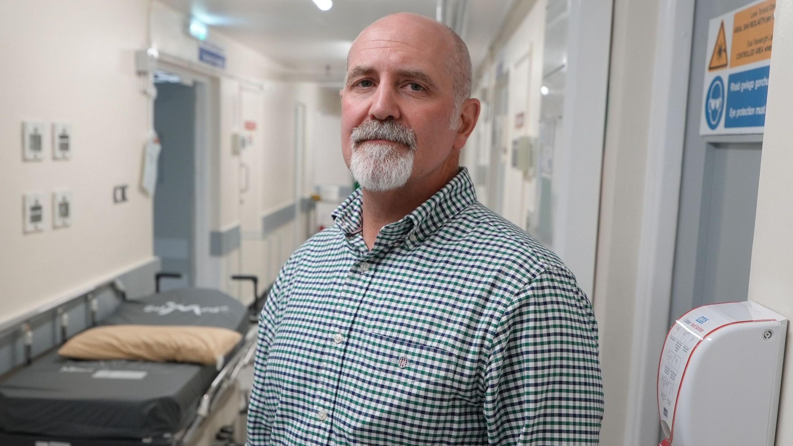 A man stands in a hospital corridor with a trolley to his left and a closed door and hand sanitiser unit mounted on the wall to his right.  He is bald with a short white goatee beard and is wearing a green, white and black gingham shirt.