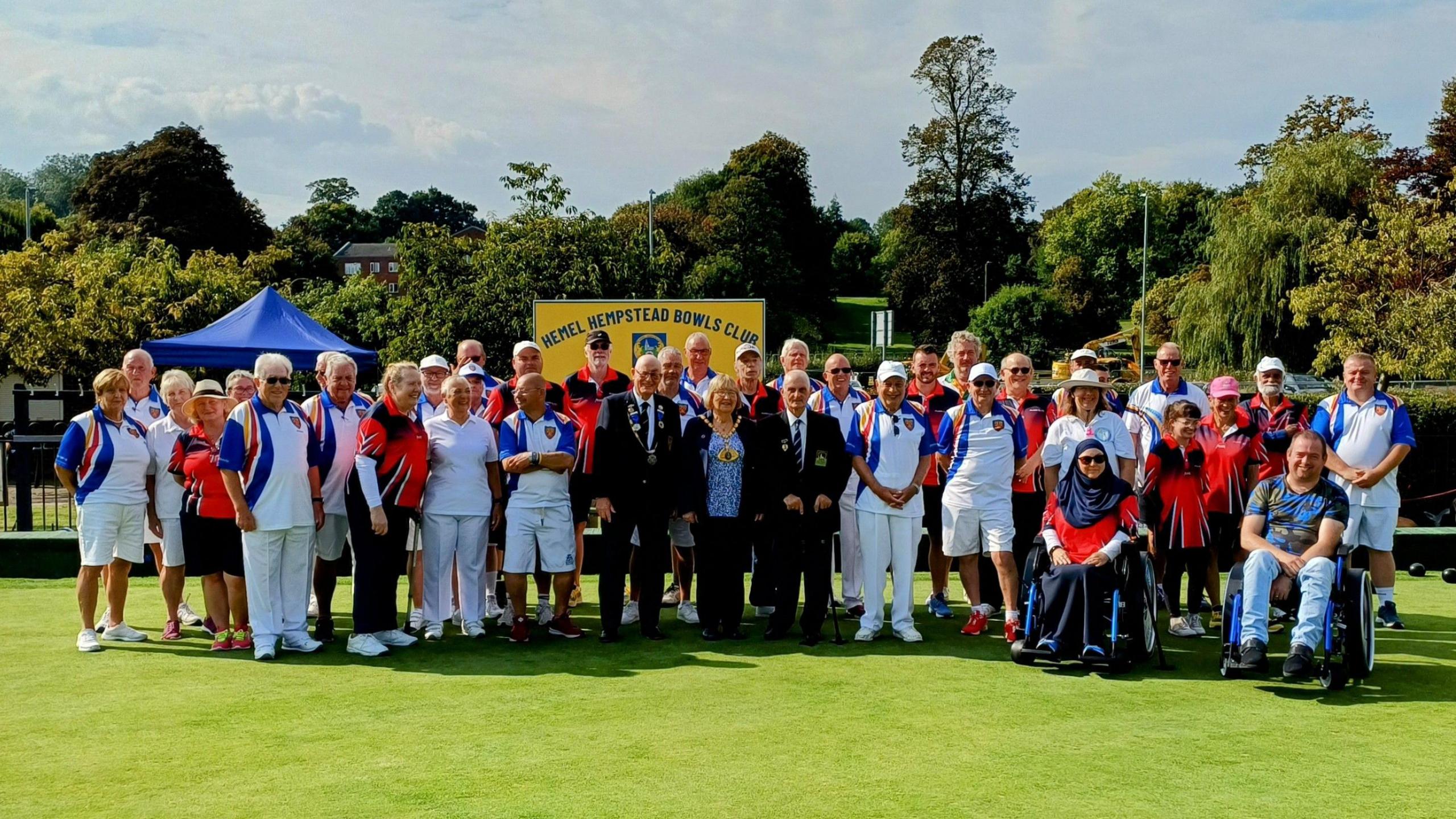 A group of people all standing on the grass of an outdoor bowling green. Some people have walking sticks, others are in wheelchairs and others are standing. Local dignitaries are also in attendance. The image is from September 2024, the last match with Disability Bowls England at Hemel Hempstead Bowls Club.