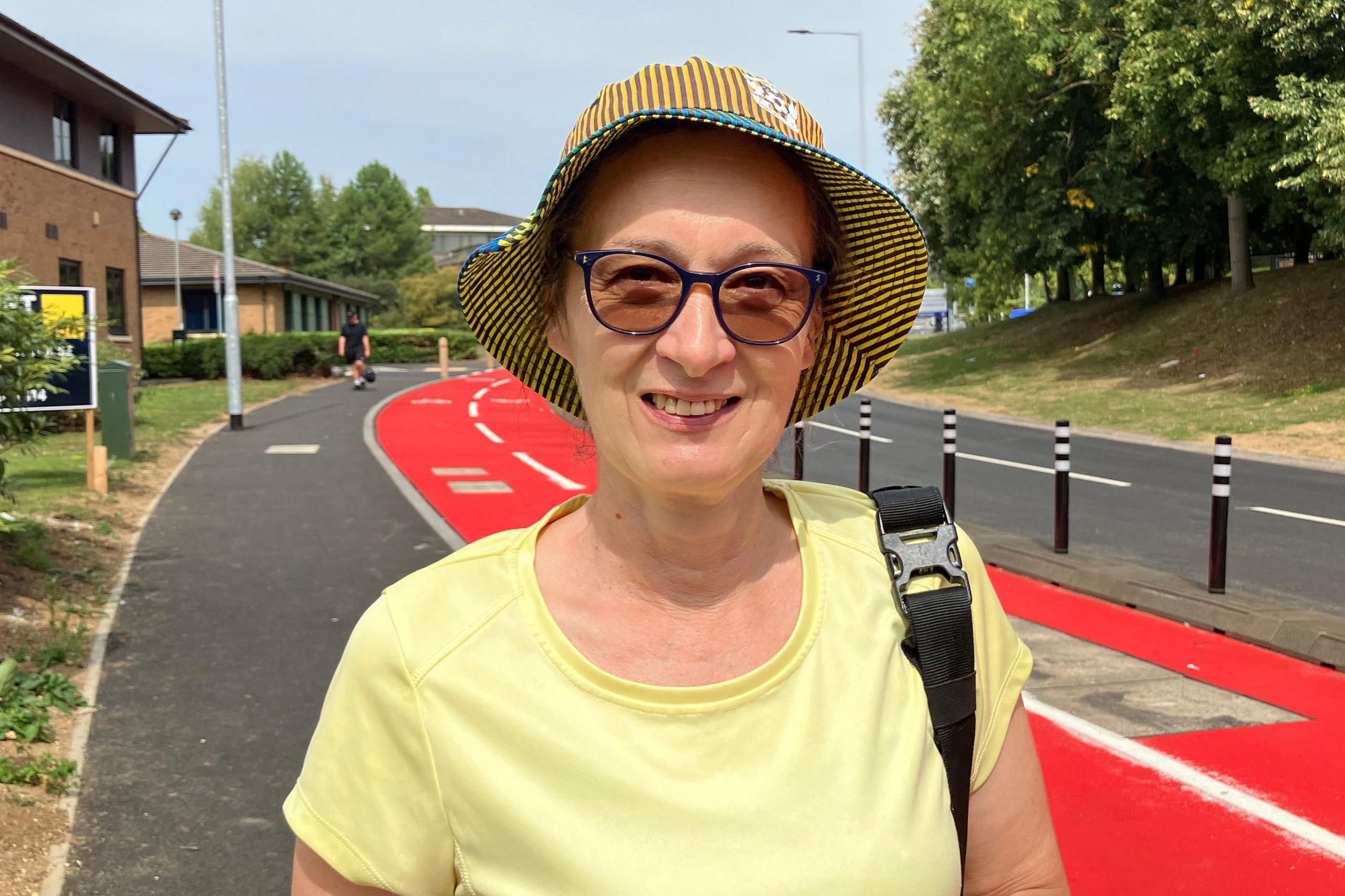 Sue wearing a yellow T-shirt and a sun hat - smiling for the camera. The red cycle path stretches behind her.