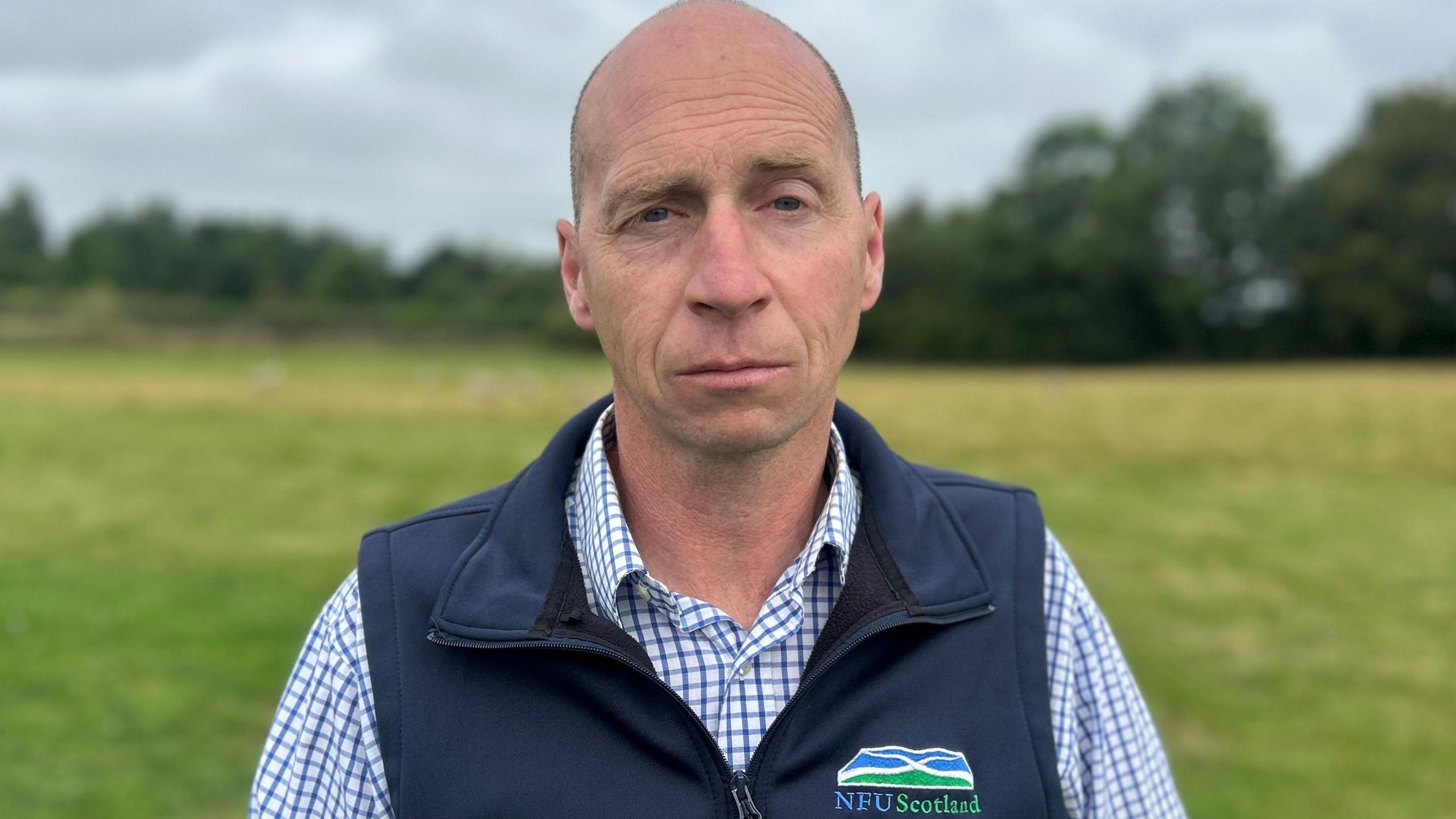 A bald man wearing a blue NFU Scotland gilet over a blue and white checked shirt. He is staring into the camera and standing in a field with a row of trees behind him.
