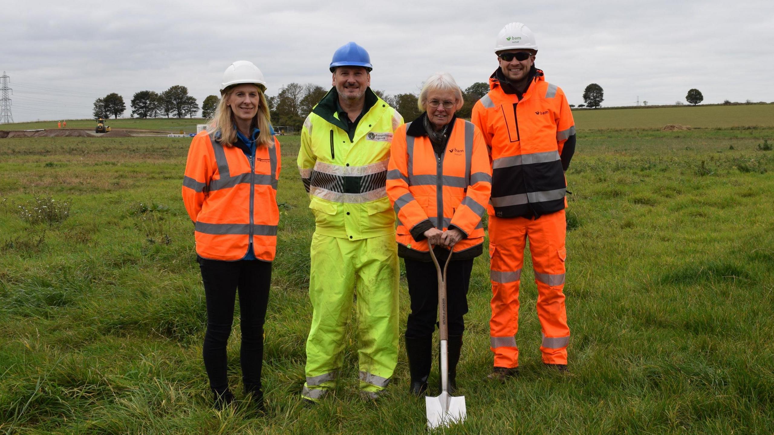 Representatives from Northumbrian Water, the EA, the Greatham Foundation and BAM Nuttall stand in full body yellow and orange hi-vis jackets in a field. Margaret Bousfield, Chair of Trustees at the Greatham Foundation, is holding a spade, blade in the grass.