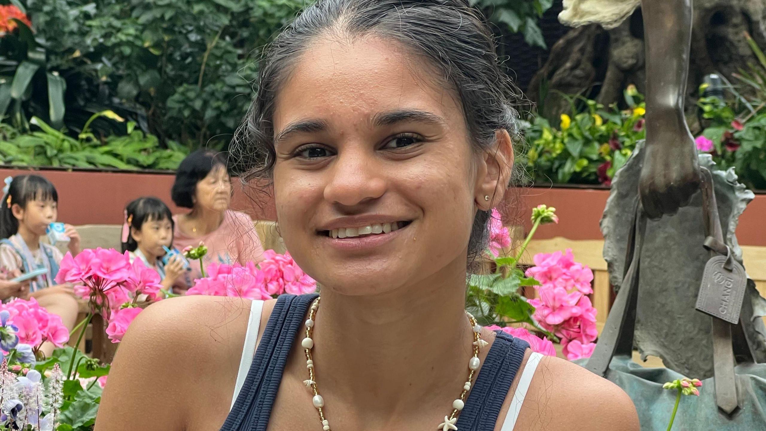 Elina Patel smiles at the camera wearing a blue T shirt and a necklace with ornamental gardens in the background