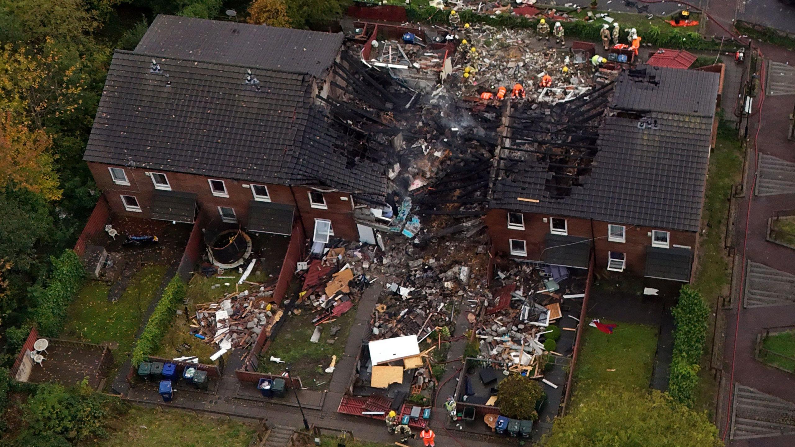 Aerial view of a terrace of six two-storey house. The middle two have been obliterated by an explosion, with the roof having collapsed and debris and rubble spread in the gardens around