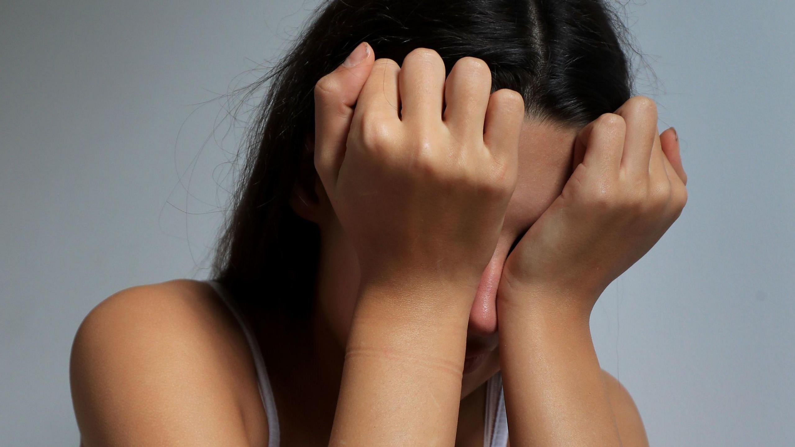 A girl with her head in her hands, rubbing her eyes. She has long dark hair and her face is covered by her hands.