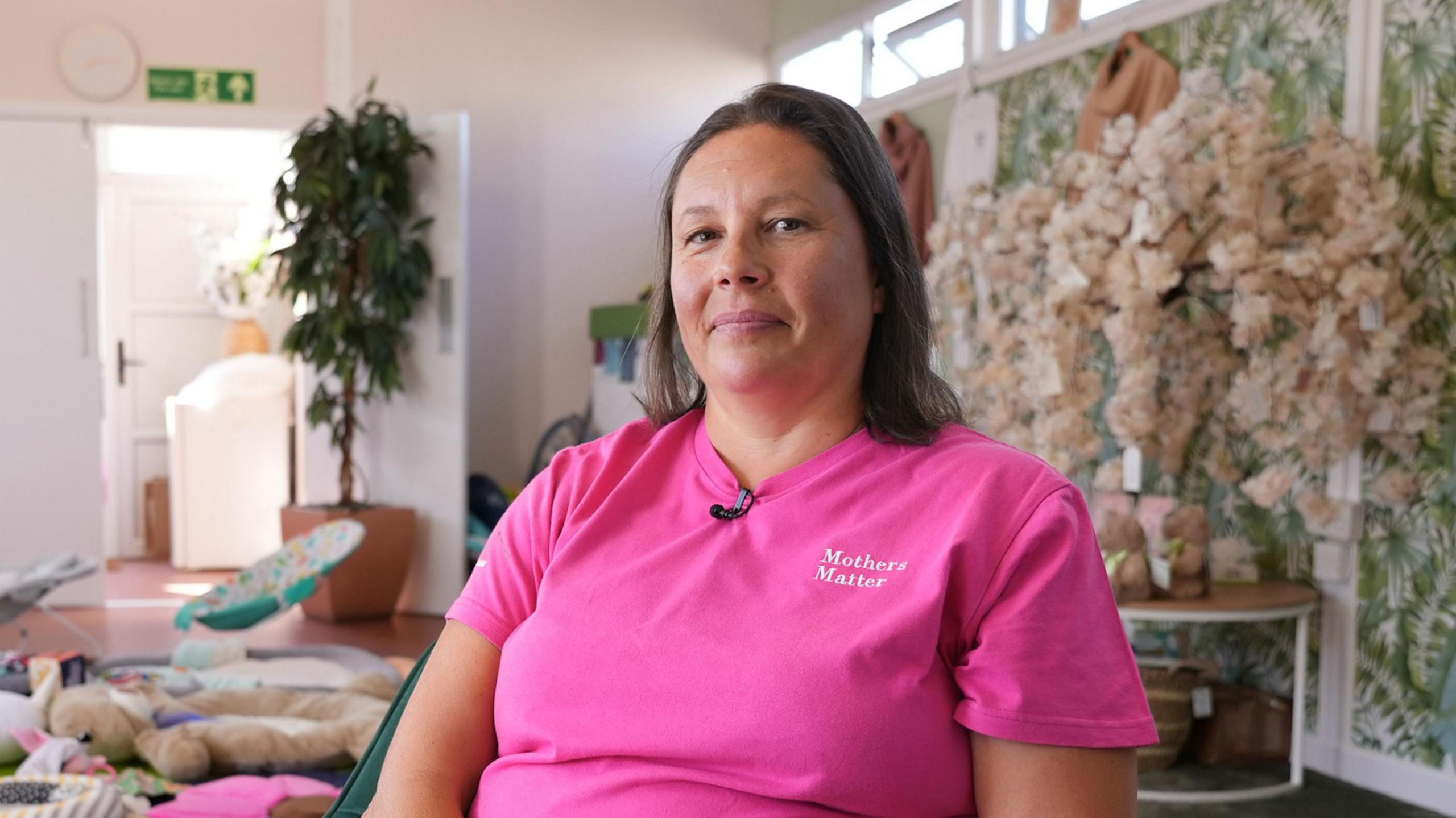 Tracy Sweetland-Hodges has shoulder length brown hair. She is wearing a bright pink t-shirt, which says 'Mothers Matter'. She is smiling and looking at the camera. On the floor in the background of the image, childrens toys can be seen on the floor.