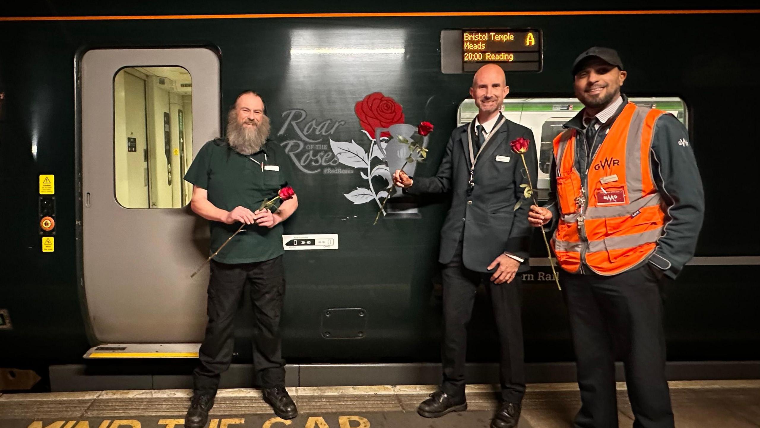Three men holding roses in front of the train. A tribute saying 'Roar for the Roses" is on the side of the train. All are smiling into the camera.