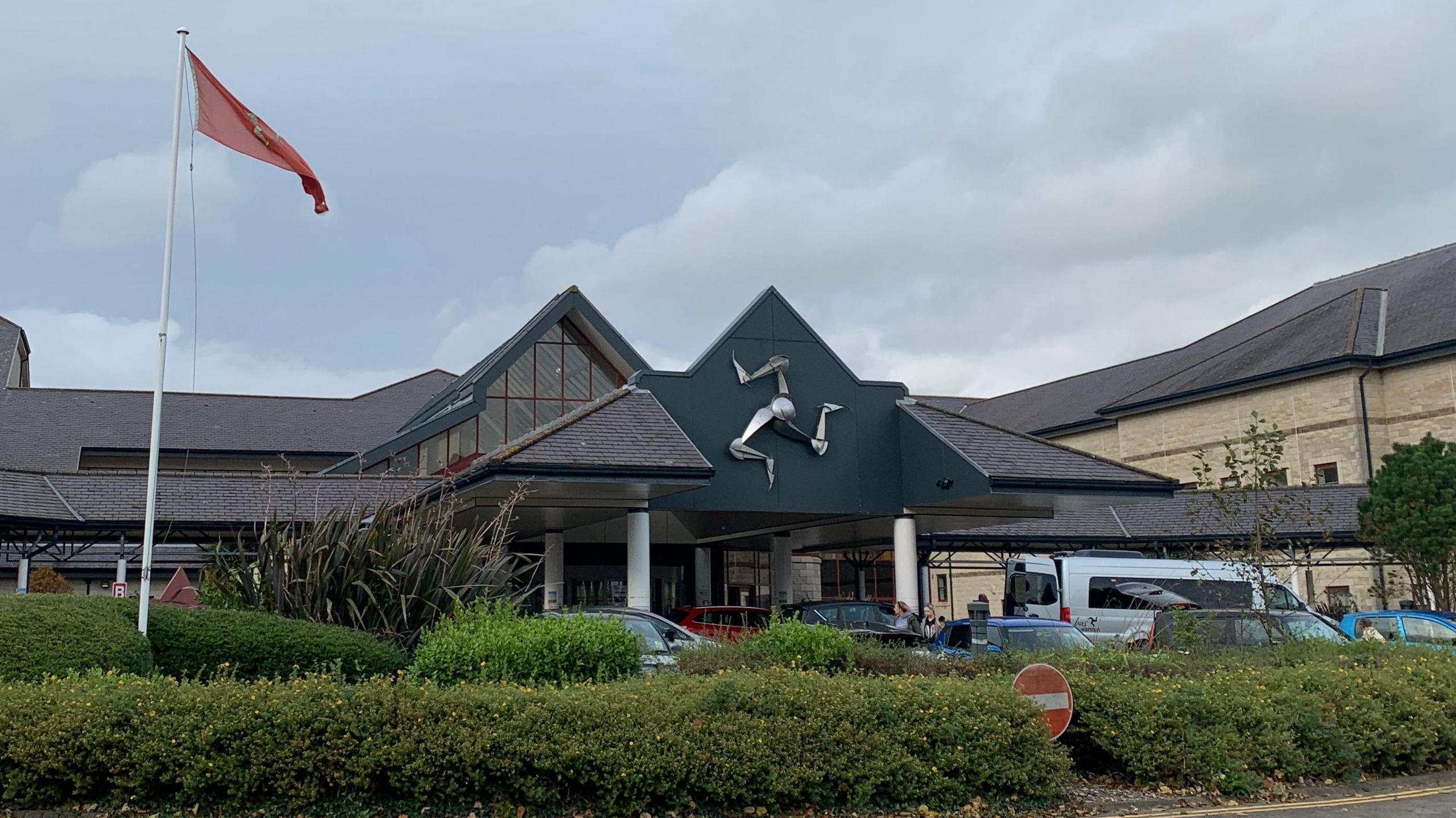 The entrance to Noble's Hospital, with the silver triskelion emblem on the front of the building and a Manx flag in front to the left above a green area of bushes and shrubbery.