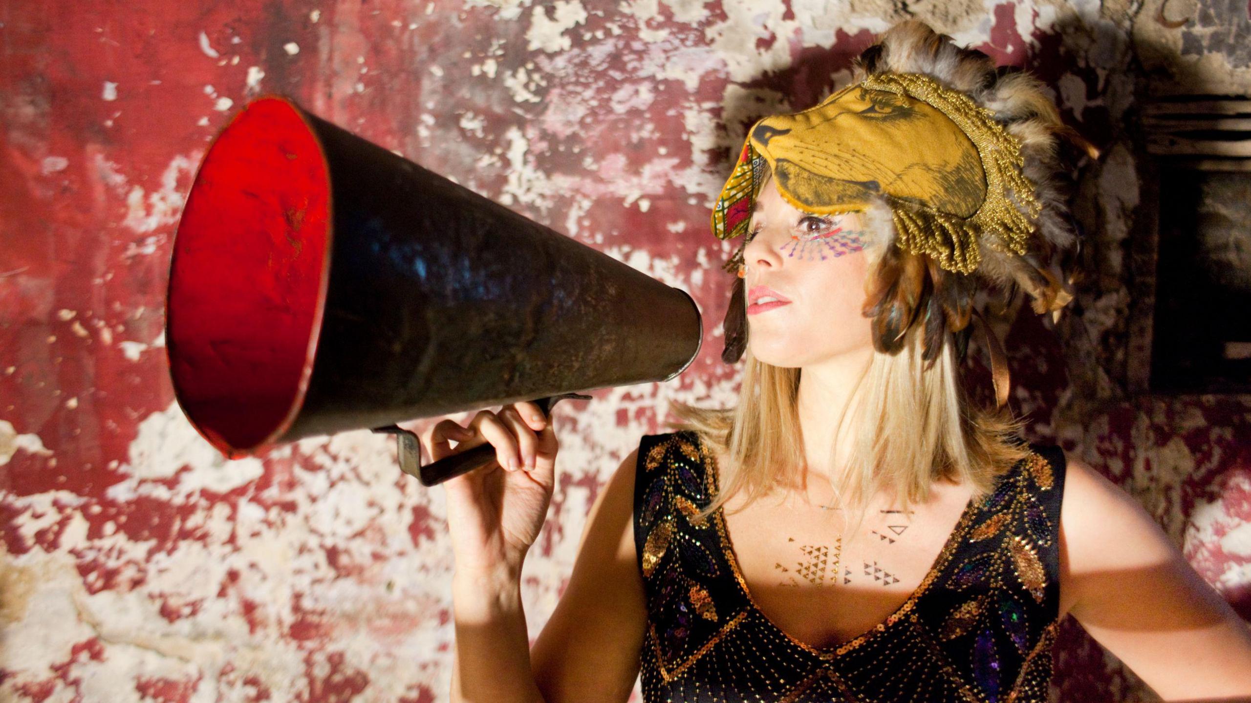 A woman with blonde hair stands in front of a worn red wall with white gaps in the paint. She wears a lion mask, which is pushed up on her head, and a sparkling beaded black and gold dress. She holds a large red speakerphone in front of her mouth and wears colourful make-up under her eyes.