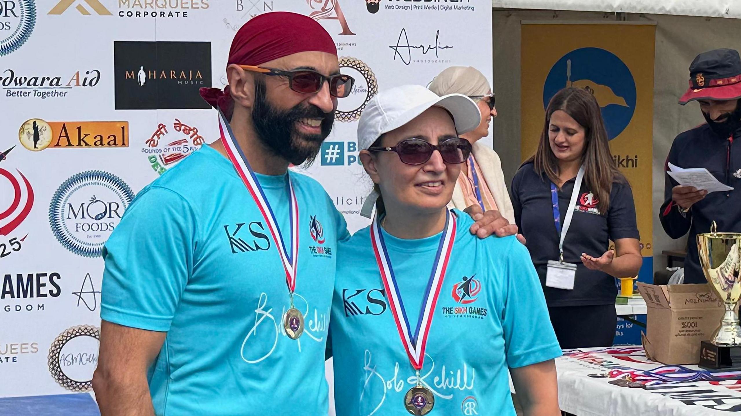 Harinder (left) wearing a neon blue top, sunglasses, a gold medal and a red head covering. He is smiling and has a black beard. Manjit (right) wears the same neon blue top, gold medal, sunglasses and a white cap. 