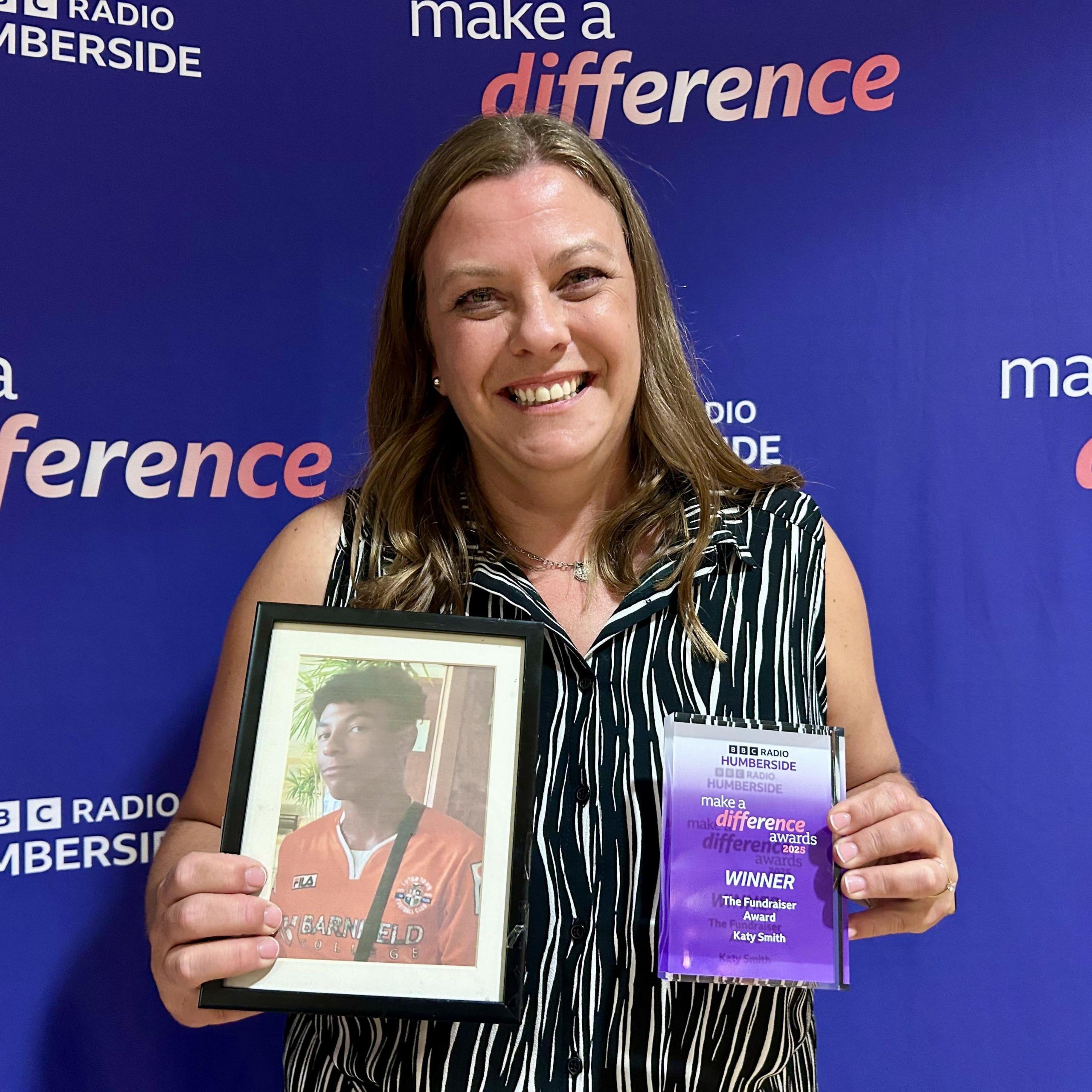Katy Smith, a woman with shoulder length, brown hair, is holding her Make A Difference award in her left hand. In her right hand, she is holding a framed photograph of her late son Ruben who is wearing a red sports top.  
