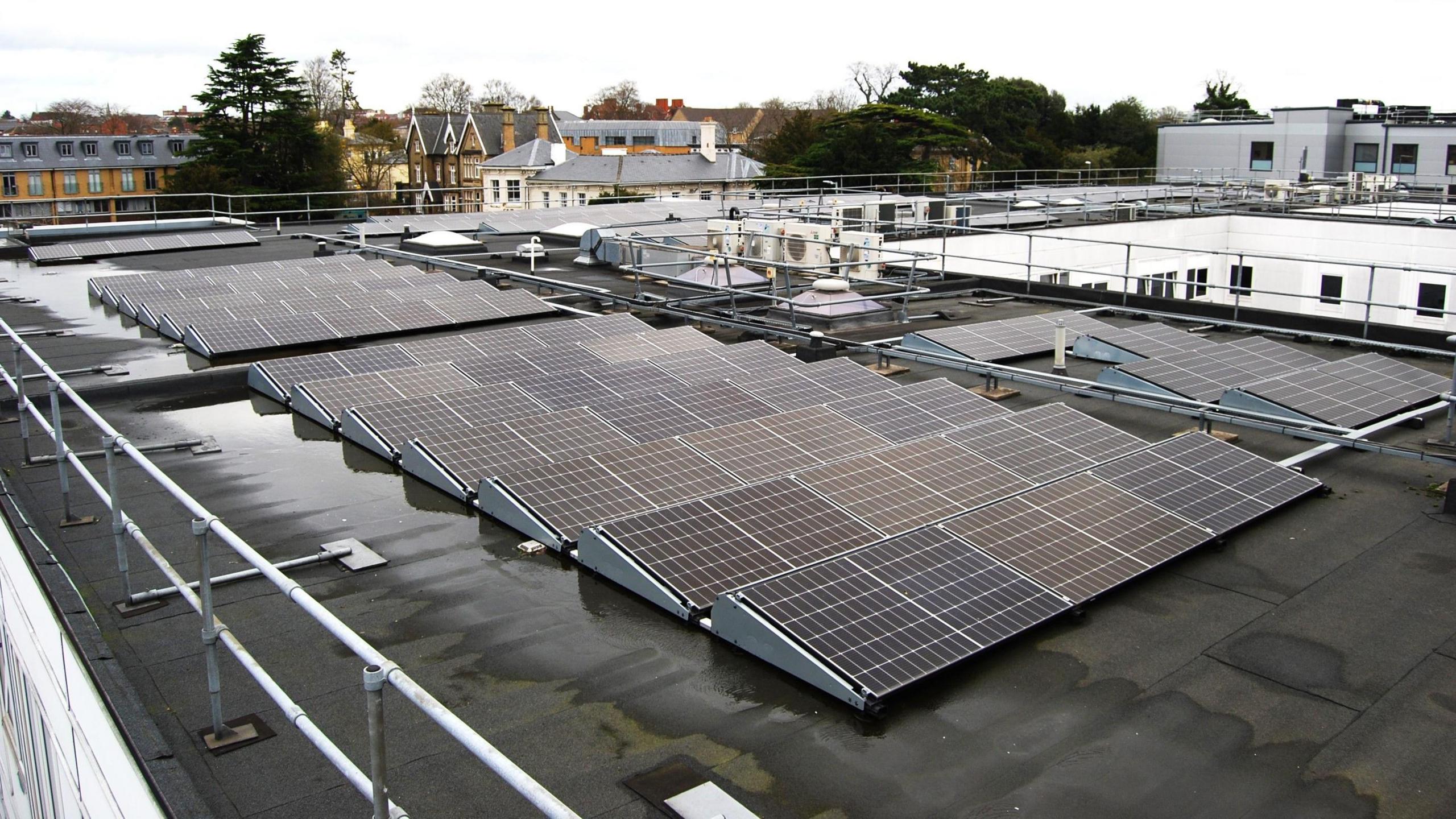 A wet and black roof of a hospital building. The roof is surrounded by white rails and on top of the roof are 13 rows of 6 solar panels. 