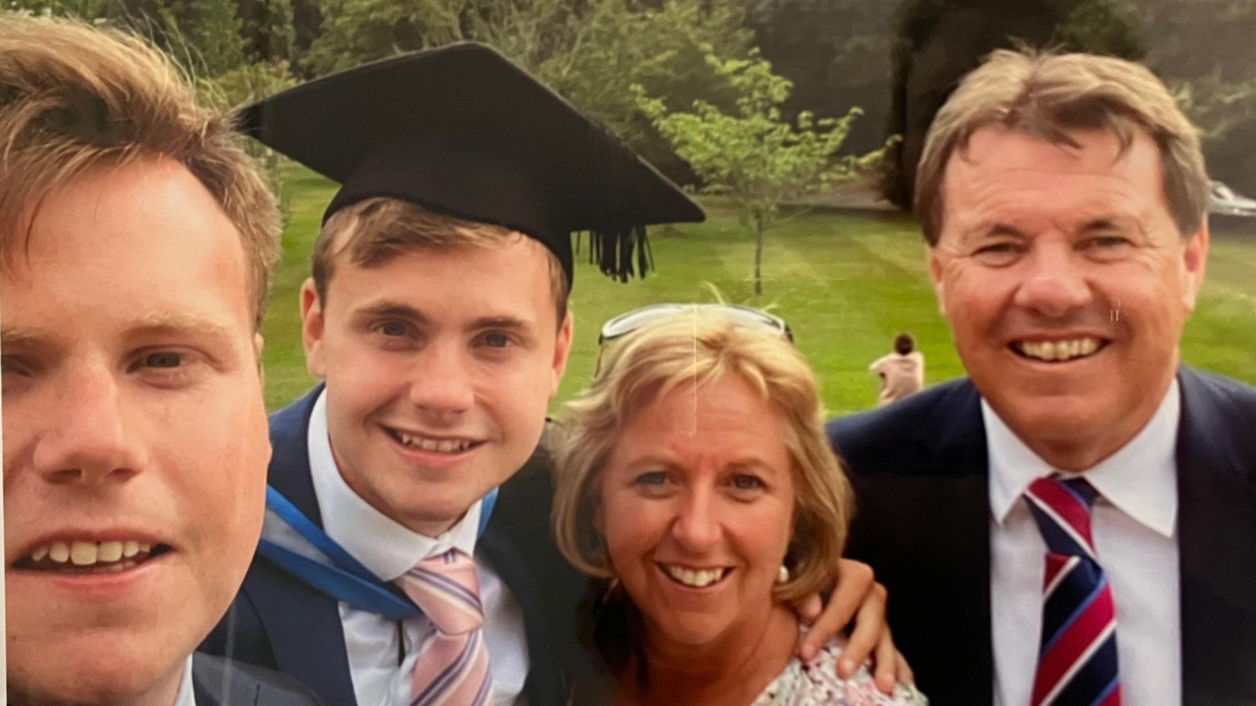 A family photo of the O'Sullivans. Jack is pictured second from the left, in a suit and his graduation cap and gown. His brother is to the left of him, taking the selfie, and he has his arm around his mother, who stands next to his father. All four family members are smiling at the camera. 