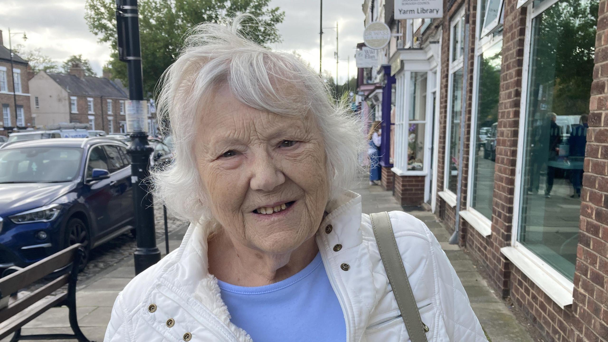 Margaret Robinson, who has white hair, wearing a blue top and a white coat, is looking at the camera. She is standing on a pavement on a high street. There is a bench to her right with parked cars.