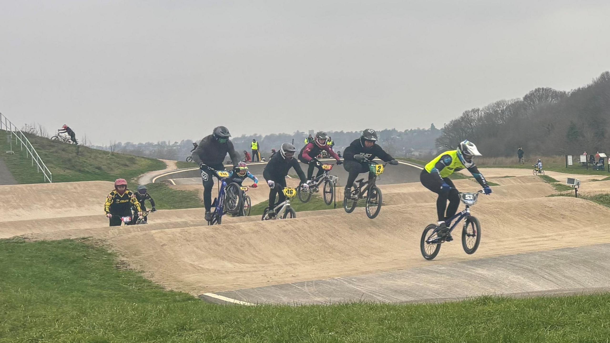 A series of BMX riders dressed in varying race gear and crash helmets cycle across the track.