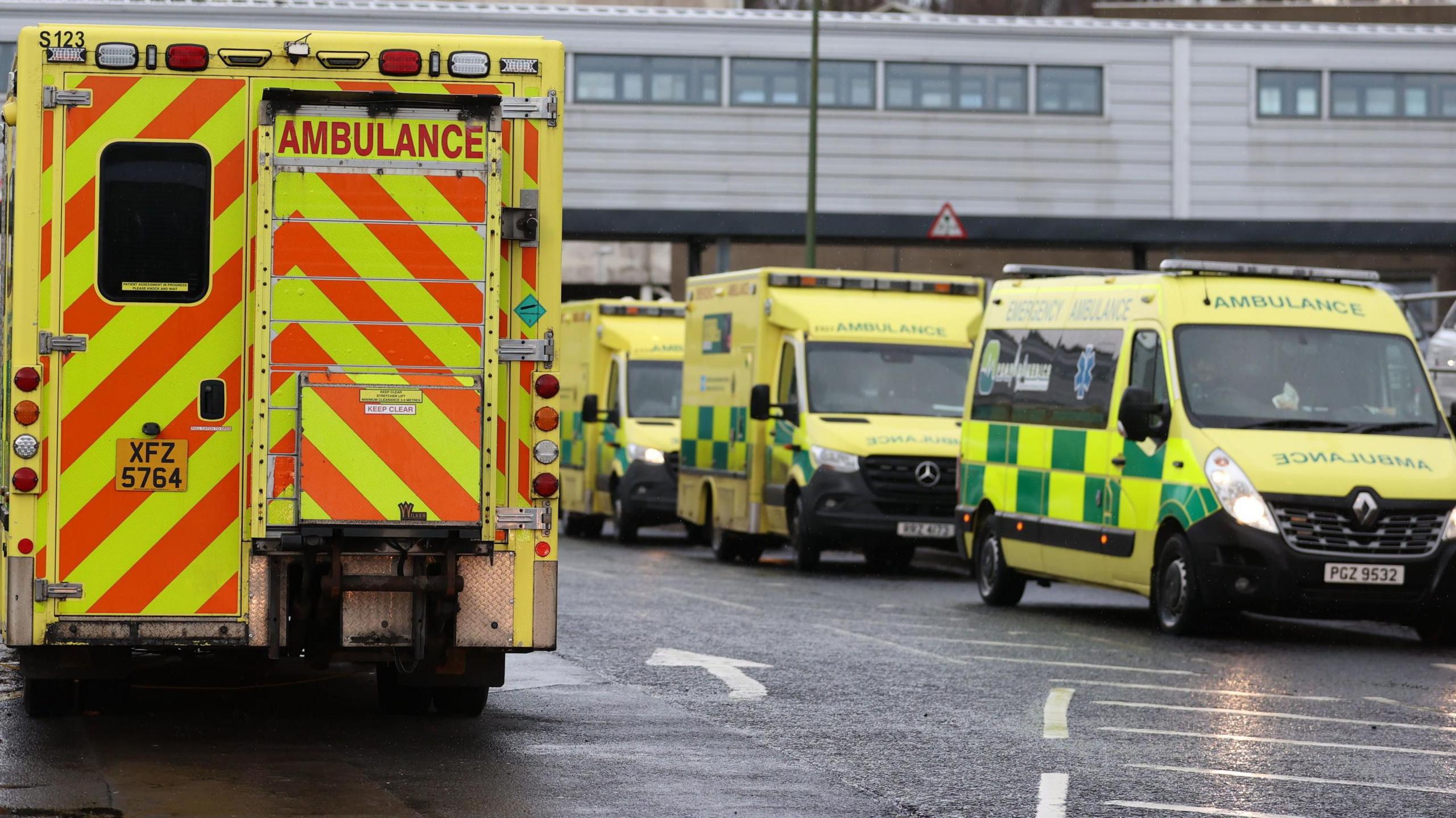 Ambulances lined up outside a hospital. Three are facing torward the camera, one has its back to the camera. They have florescent livery. 