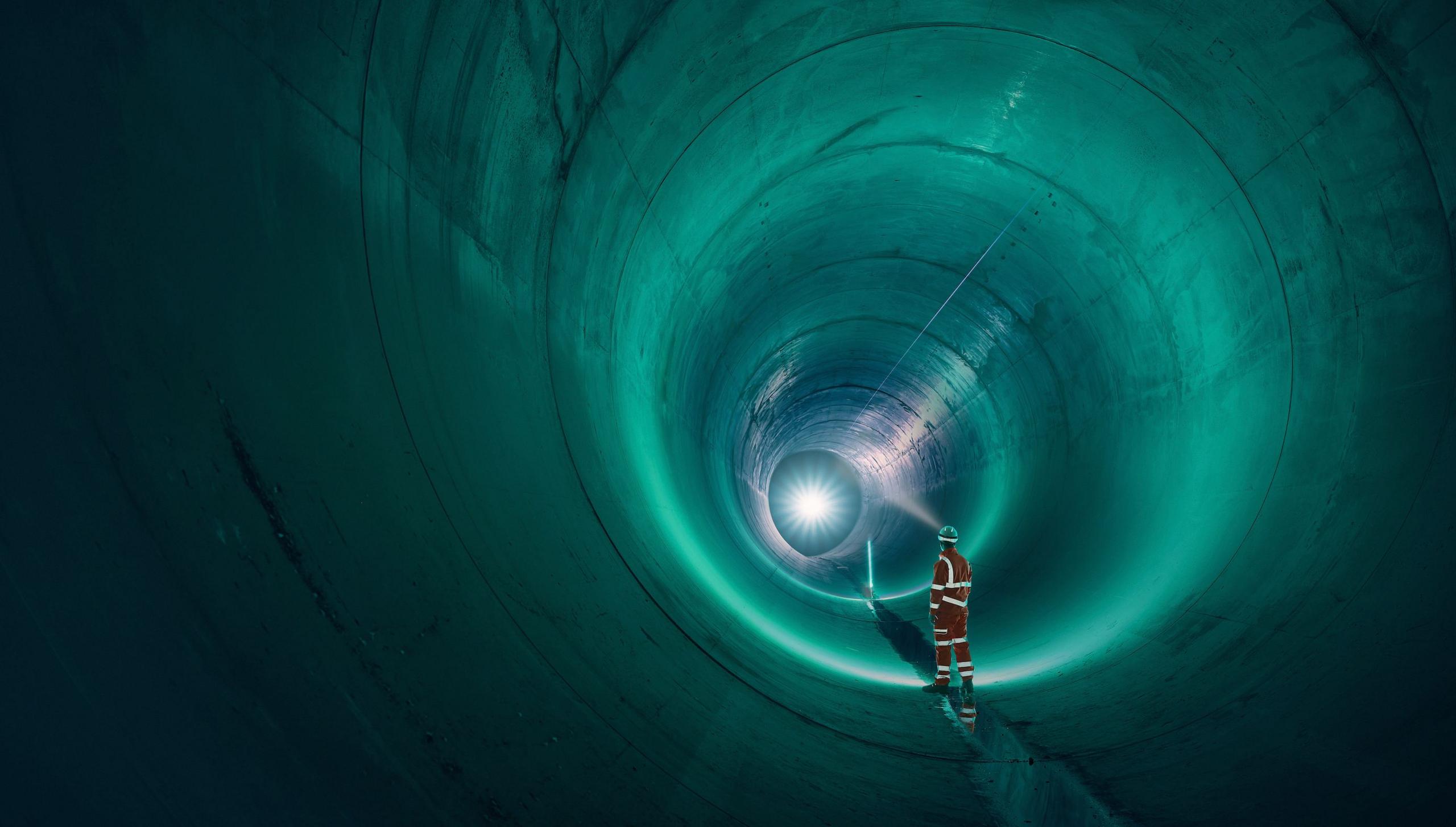 A worker in a high-visibility orange suit and helmet stands inside the massive, curving tunnel of the Thames Tideway Tunnel, illuminated by greenish-blue light. The tunnel extends into the distance, with a bright light shining at the far end.