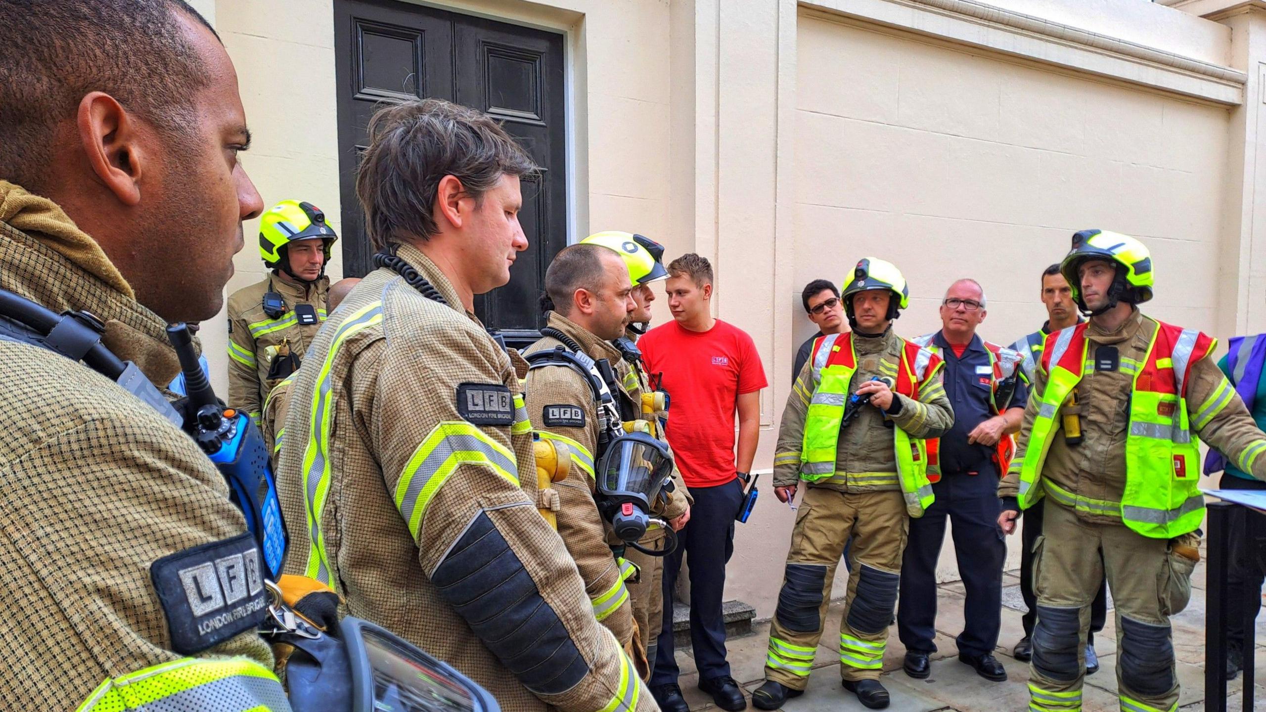 A group of at least 12 firefighters stood in a group listening to someone speak. They are all wearing LFB uniforms which are brown, with yellow hi viz on their arms, backs and trousers, some are also wearing hi viz jackets and bright yellow helmets. 