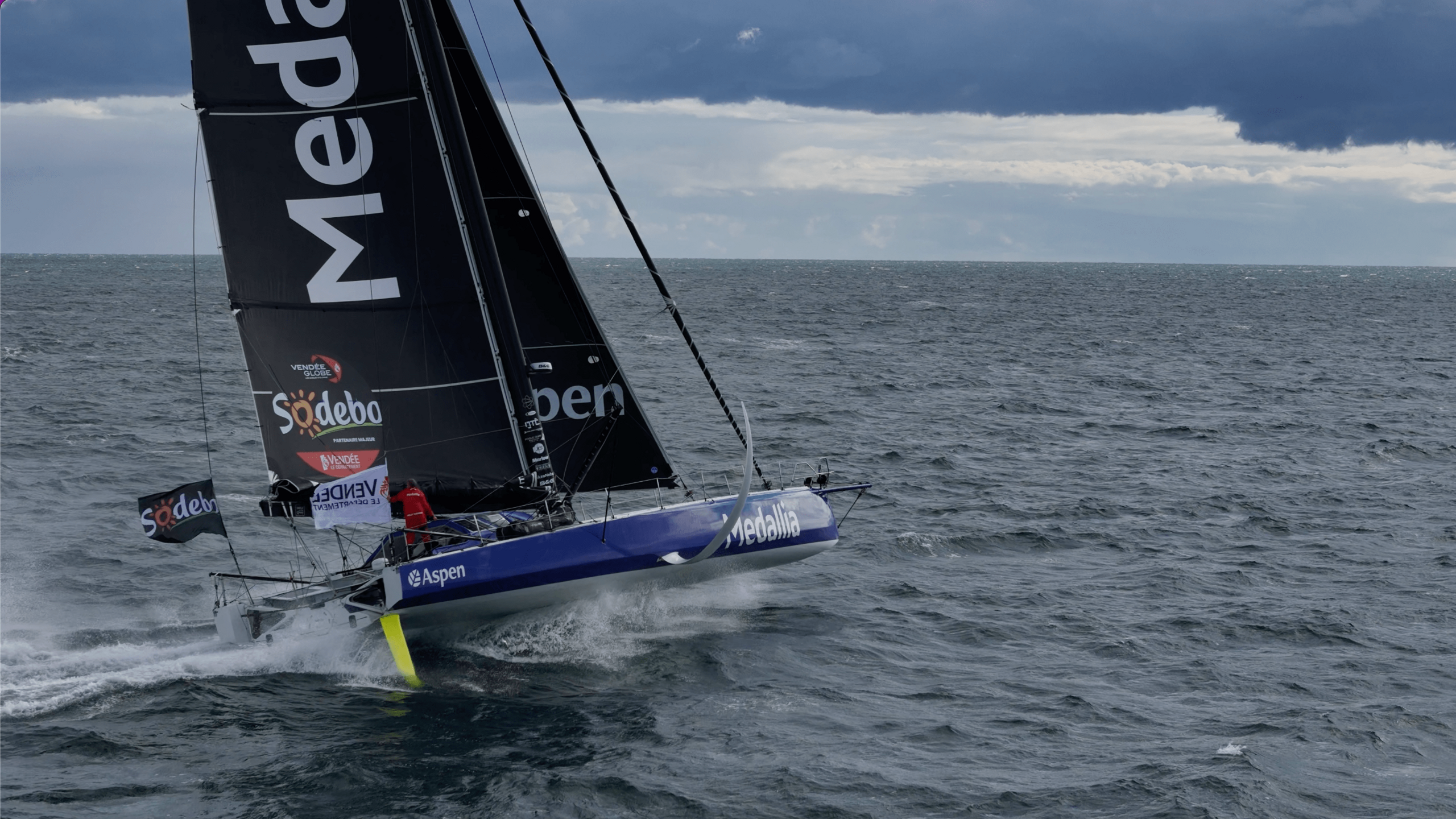 A blue sailing boat with a large black sail is travelling through water at speed. The sky is grey.