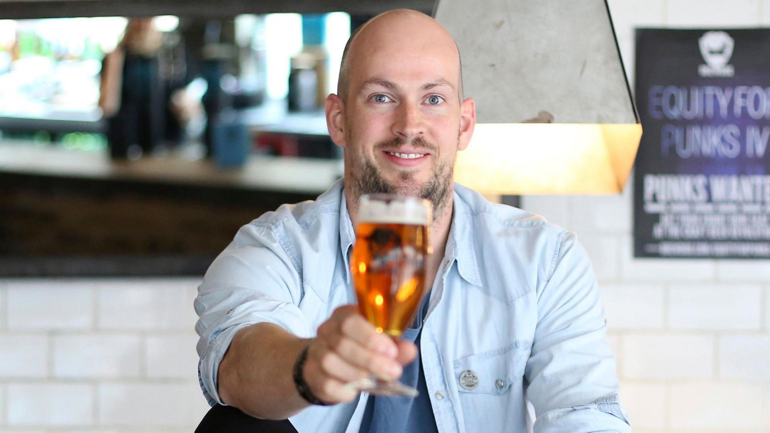 A smiling bald, bearded man wearing a light blue shirt - with the sleeves rolled up - and a light blue t-shirt raises a full beer glass with his right hand. He has been photographed in a pub setting, with a white wall and a bar visible in the blurry background.