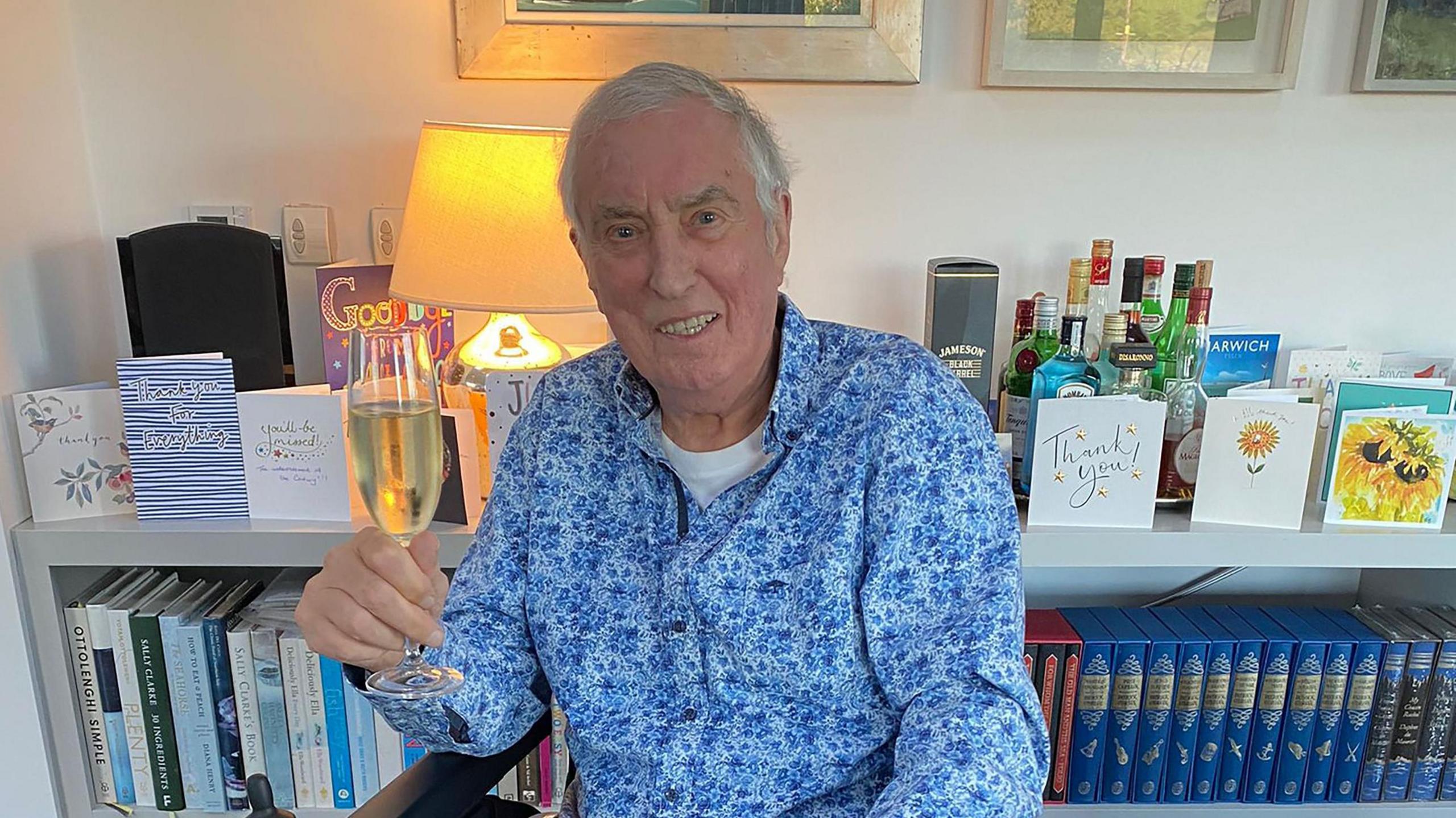 Johnnie Walker, pictured in a wheelchair holding a glass of champagne after his last programme. He has a floral blue shirt on and slippers. He is at home and behind him you can see cards on a bookshelf, these are some of the cards listeners sent to him. 