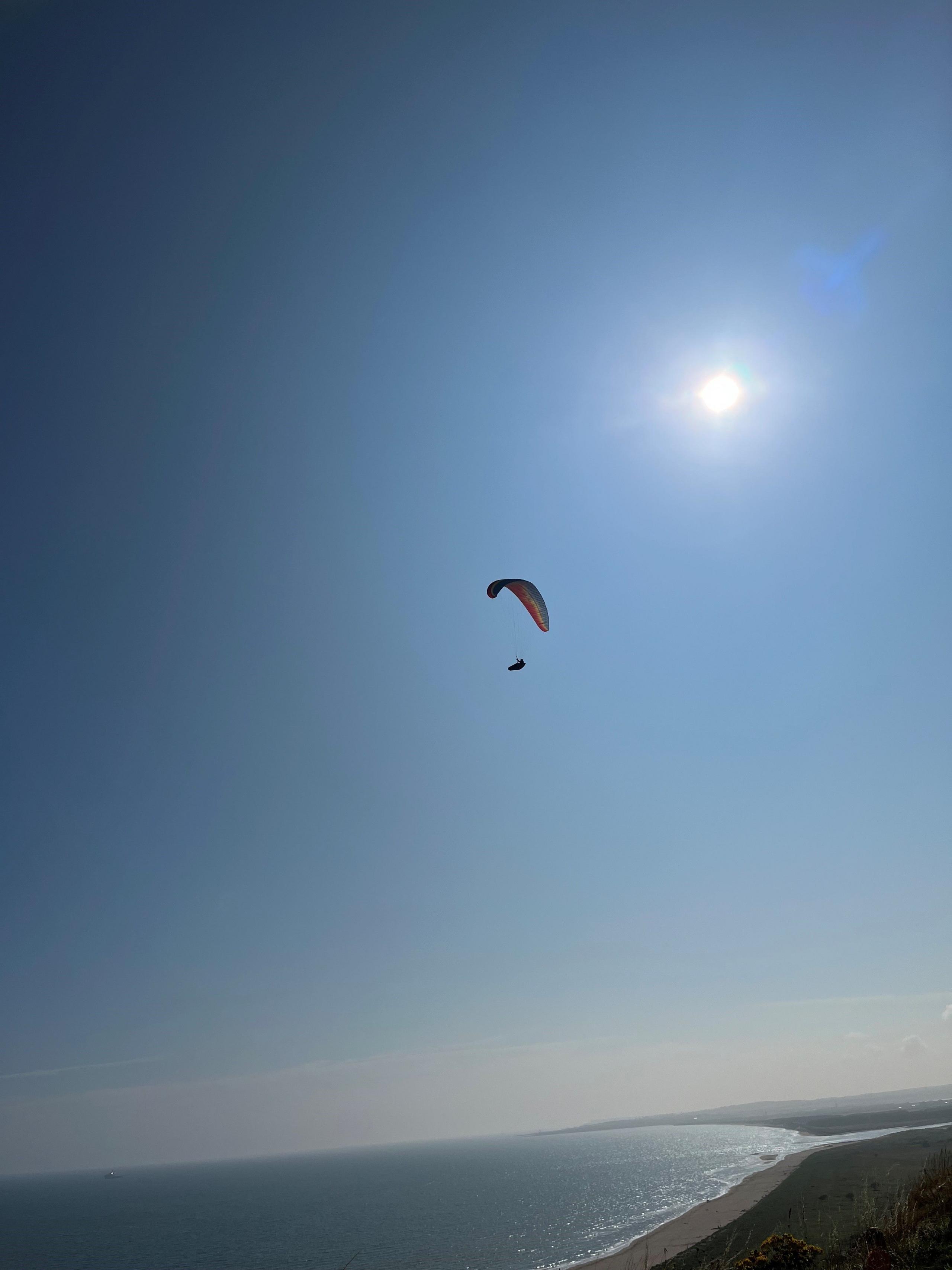 A unidentified person windsurfing over a beach in Aberdeenshire. The sun is high in the sky in background.