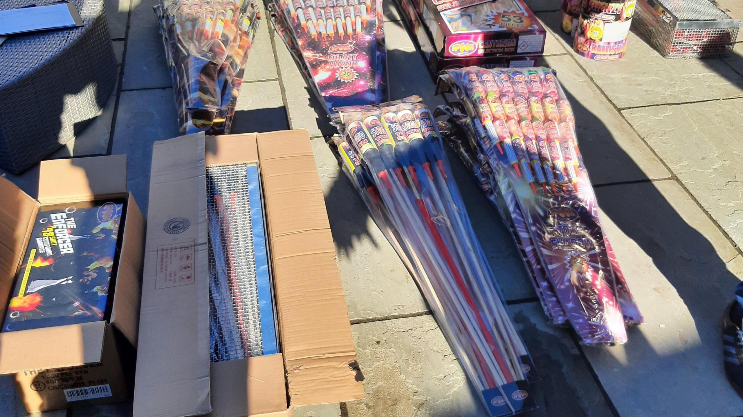 Piles of fireworks are laid out on a paved area. Two open cardboard boxes containing fireworks are on the left. Rocket fireworks on sticks are laid out in four piles. 