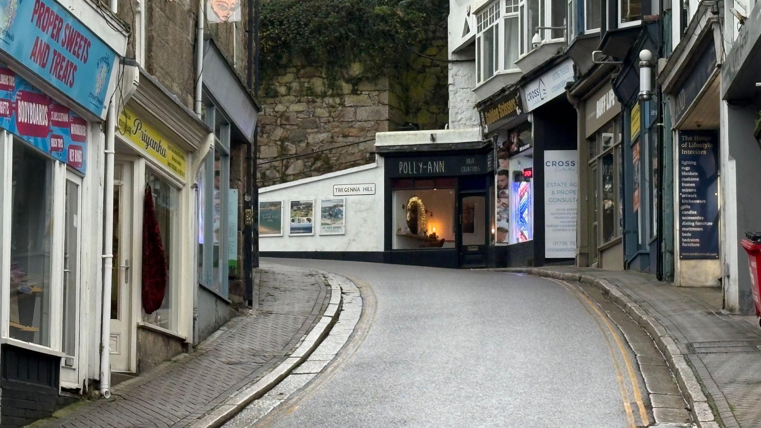 The narrow lane leads up hill and turns sharply to the left. There are small shops lining it and no people are in sight.