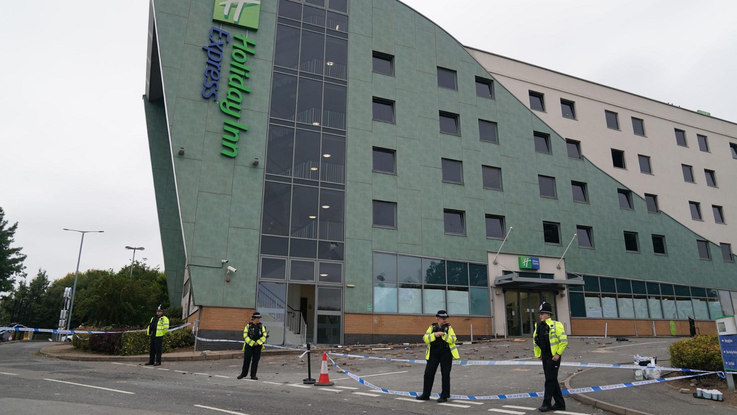 Holiday Inn Express building with four police officers outside with police tape and orange and white cones visible. 