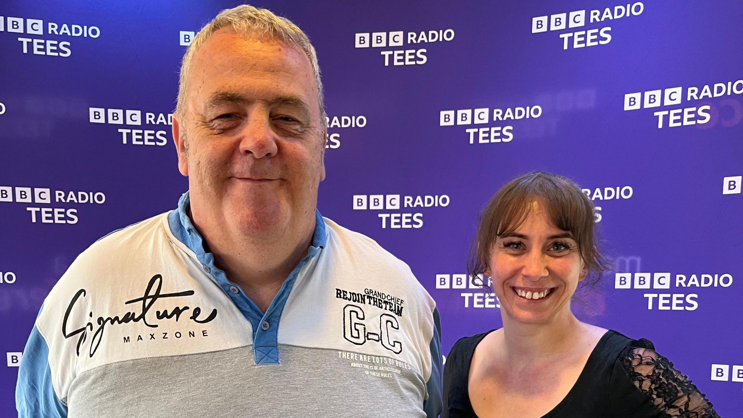Ian and Nancy are standing in front of a purple BBC Radio Tees branded background and smiling. Ian has short white hair and Nancy has brown hair.