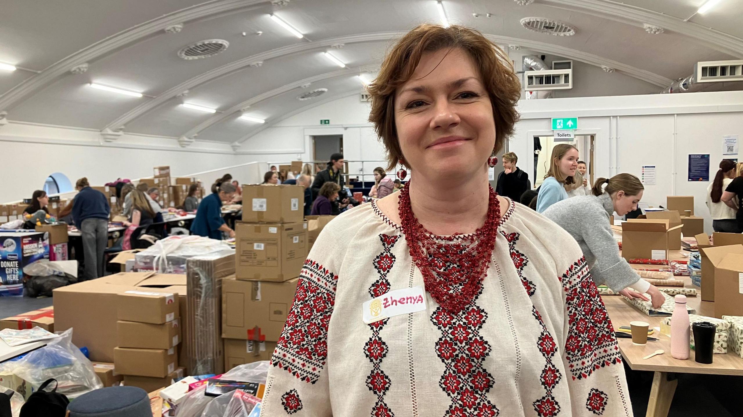 A woman wearing a festive jumper stands inside a large room in Swindon where a wrapathon is taking place in aid of Ukrainian children. Behind her are several boxes of gifts, and volunteers can be seen working on tables