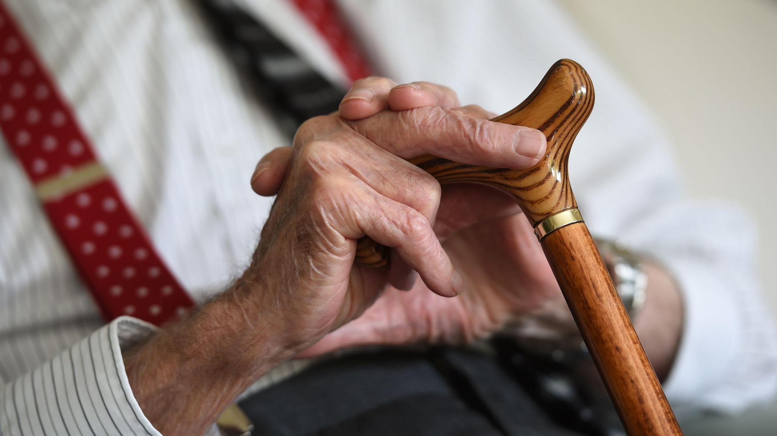 A close-up image of a man sat down, holding a walking stick - his hands are visible, as well as a brown stick, and part of his upper body. He is wearing a white shirt and red braces.