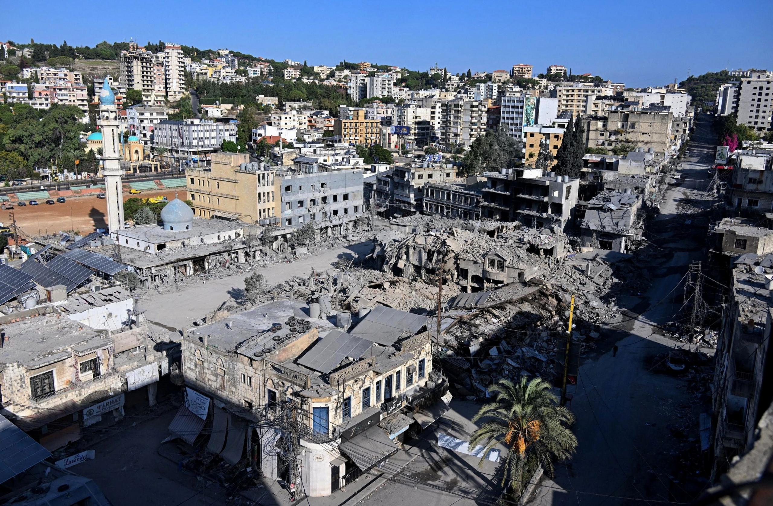 The destroyed market in Nabatieh. Grey debris from a pulverised structure covers a central square, which is lined by buildings with their windows blown out. High-rise buildings stand on the hills in the background. 