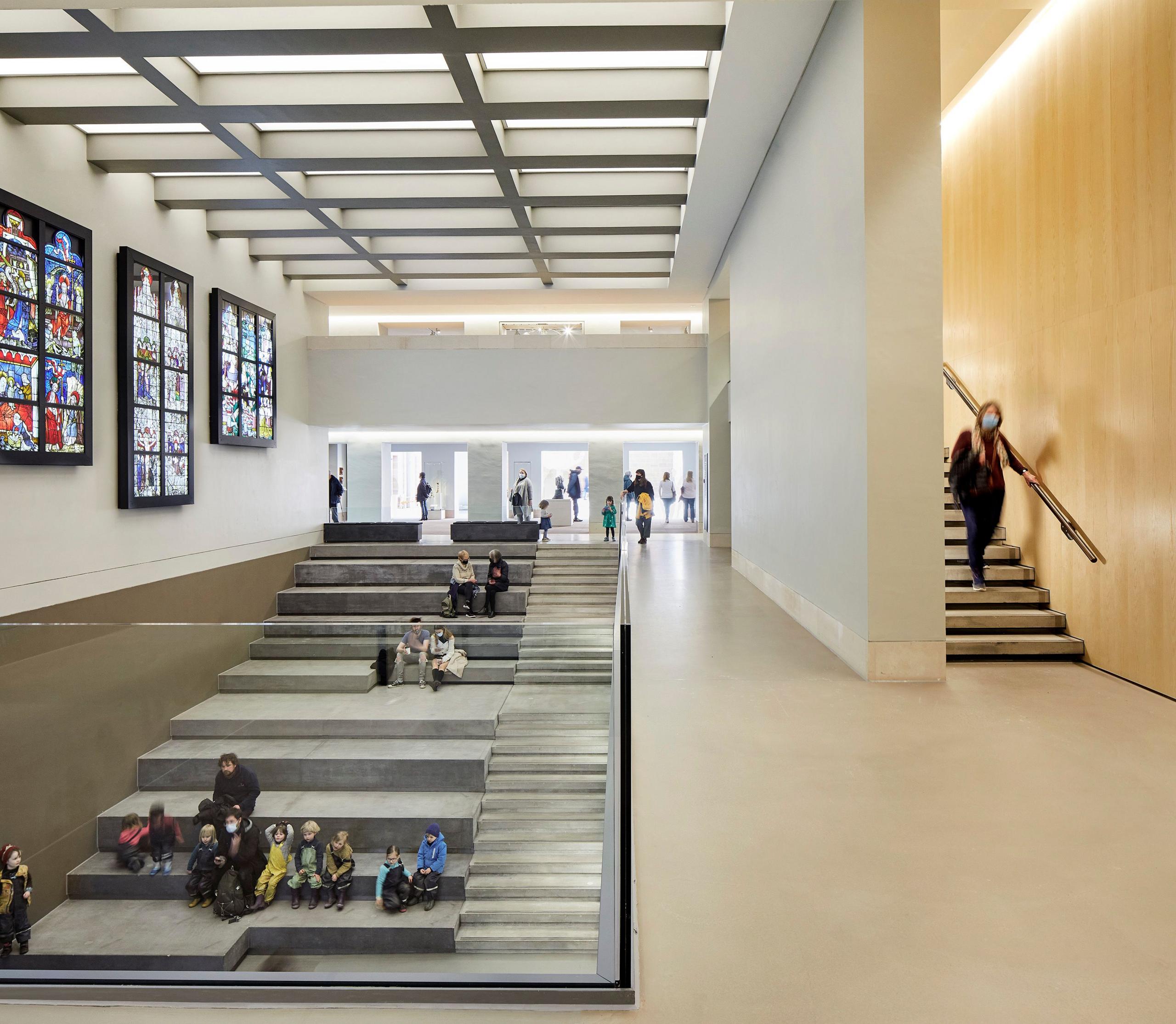 A stairway extends the steps to create a tiered seating area. People sit on these steps below a display of colourful stained glass on the wall above.