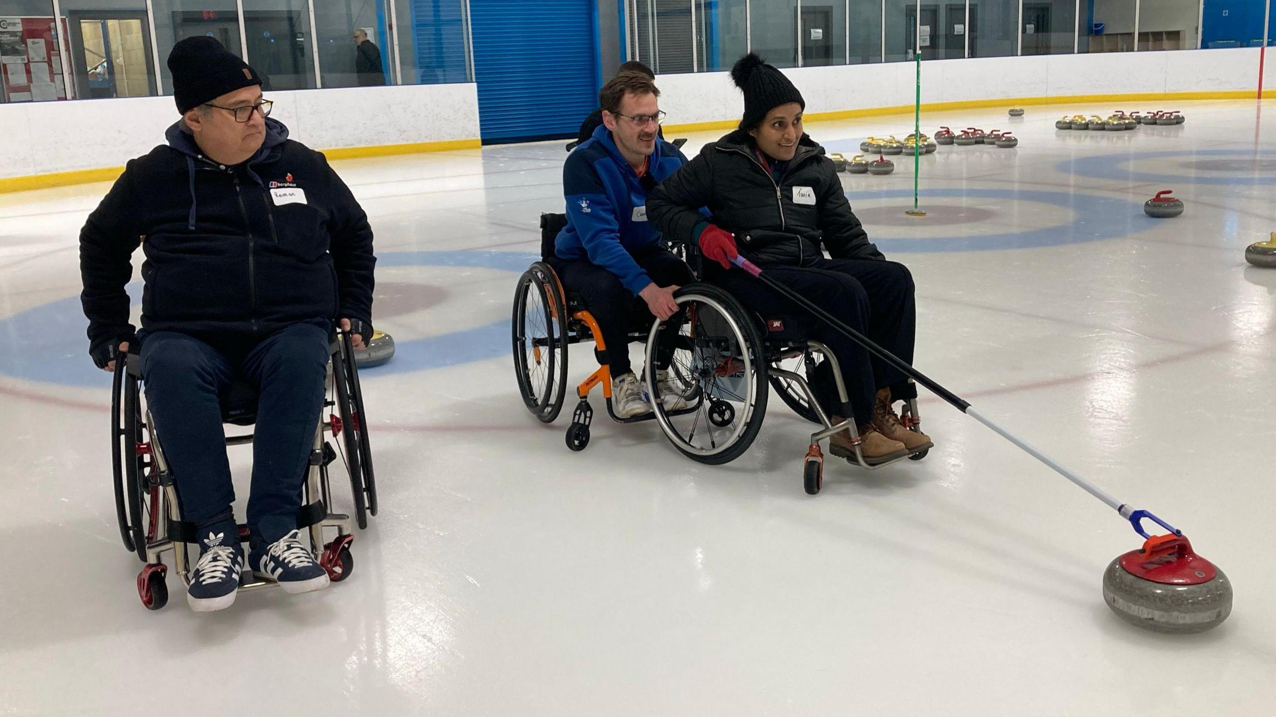 People in wheelchairs trying curling in Cambridge. A woman in a wheelchair is being guided by a man, also in a chair. She has a curling stone on a long pole. Another man in a wheelchair is watching from the side