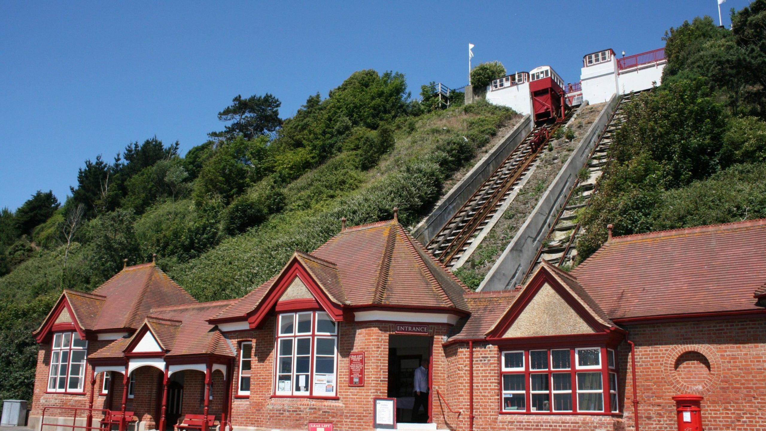 The Leas Lift in Folkestone, with the Victorian red brick ticket hall at beach level and white stone buildings at the upper promenade level. A red carriage is at the top level on the left hand set of rails.