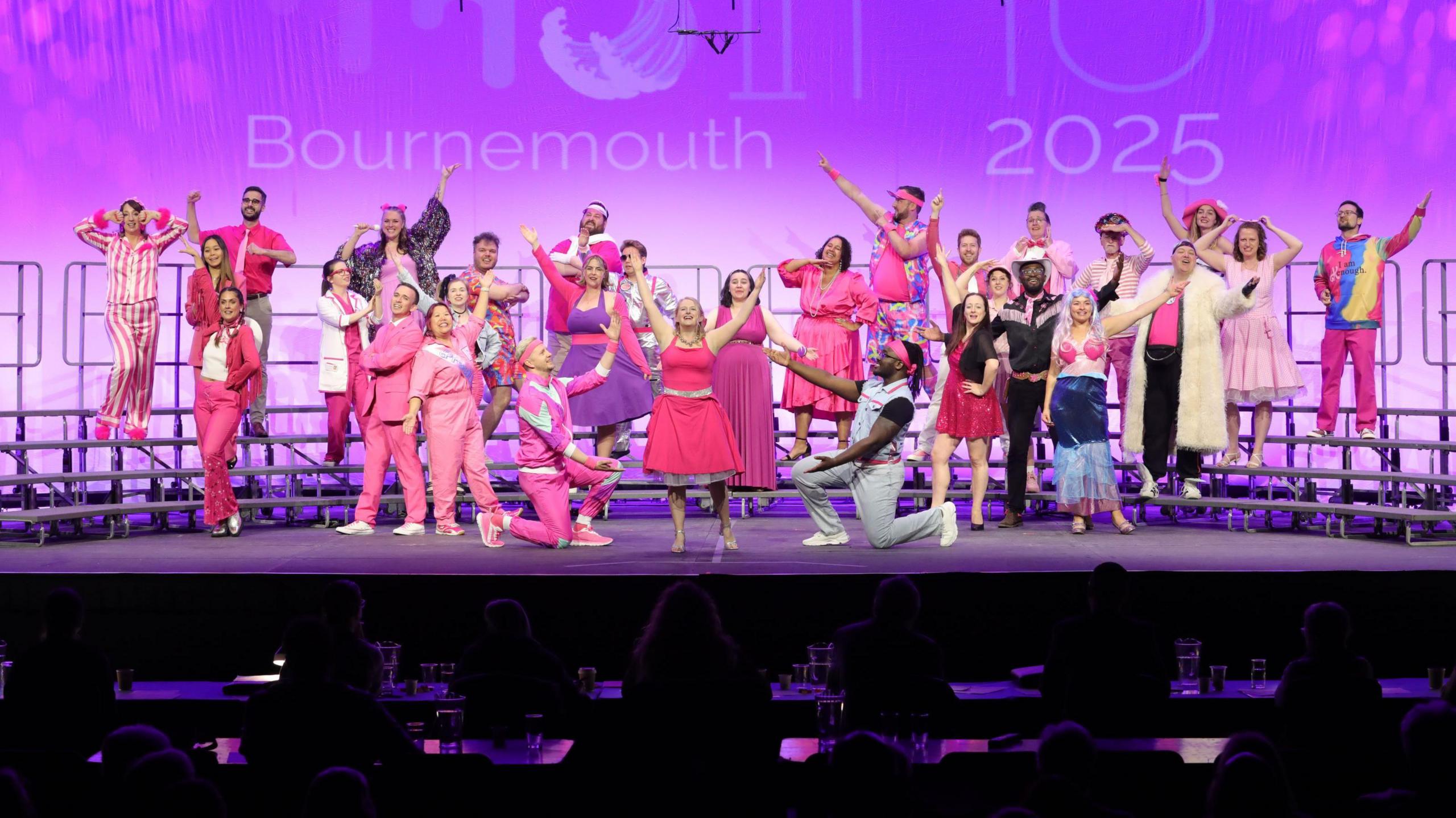 A large group of singers from Bristol A Cappella choir sing on stage at the choir awards in Bournemouth. Many of the choir are wearing pink and they are showing passionate facial expressions and poses