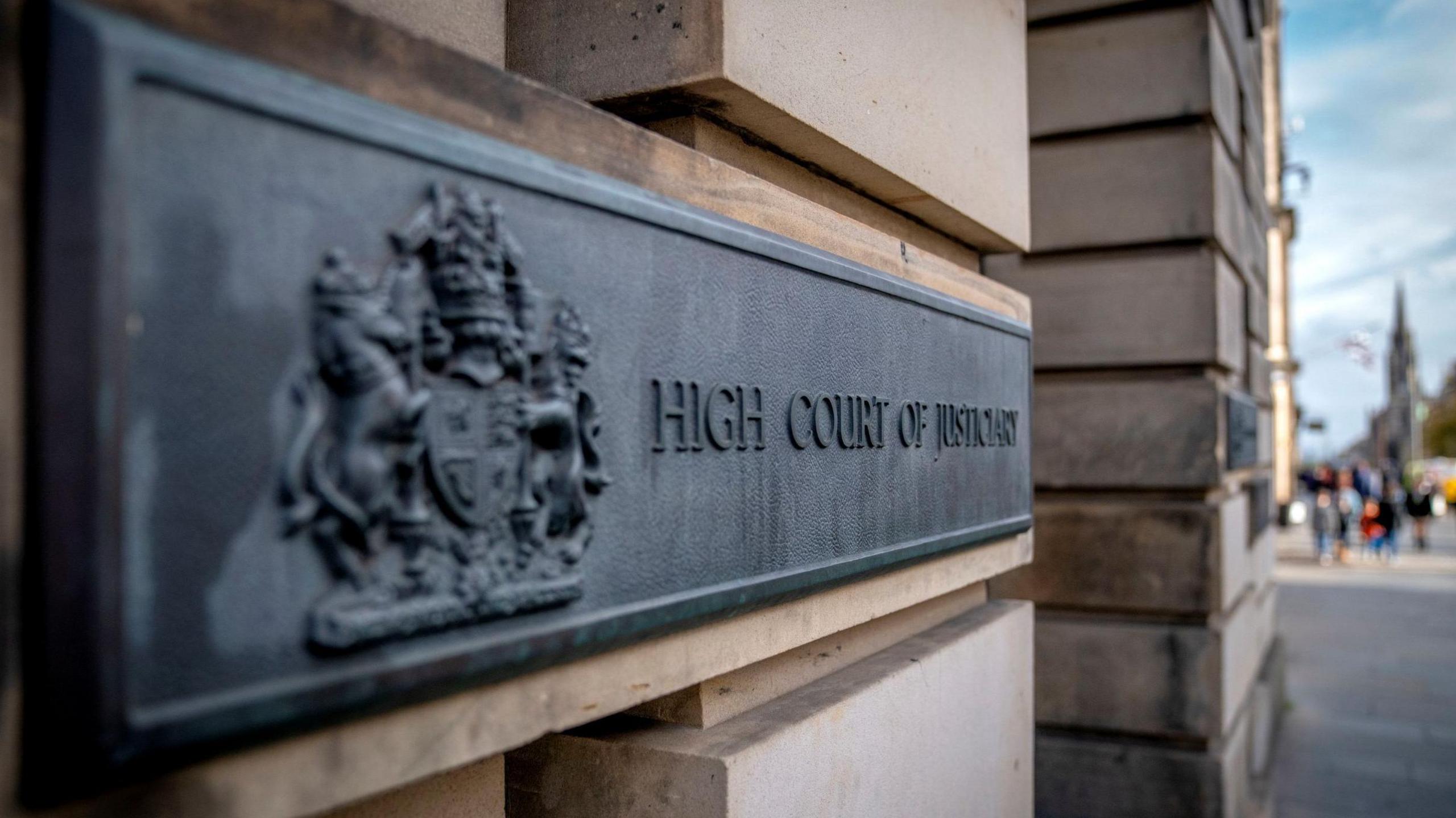 A dark grey metal sign on the sandstone block wall of the High Court in Edinburgh. The sign has a royal crest and the words: "High Court of Justiciary".
