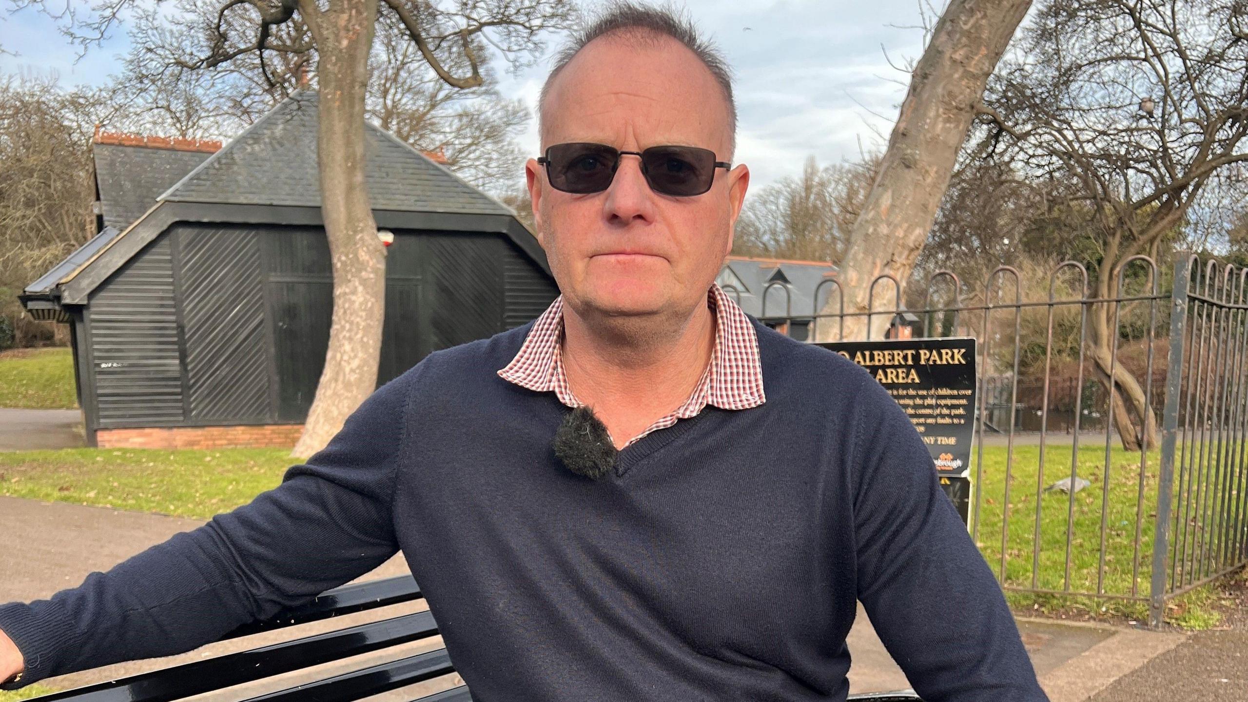 Mark Knight is sitting on a park bench wearing a grey jumper and dark glasses. There is a grey building in the background and park railings. 