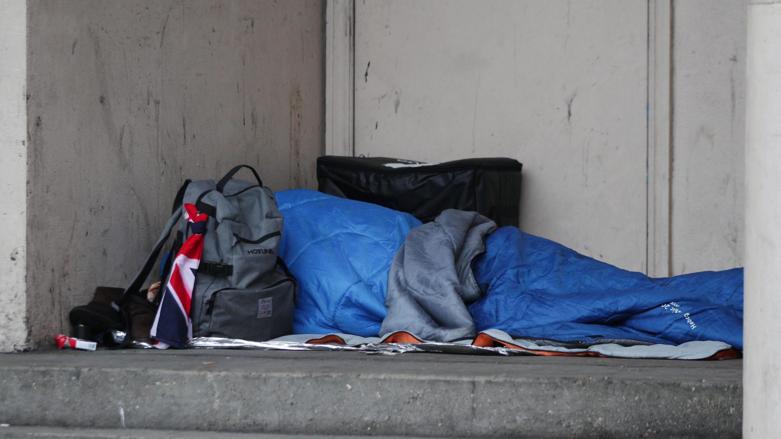 A blue tent depciting a rough sleeper with a grey rucksack nearby with part of a Union Jack flag showing attatched.