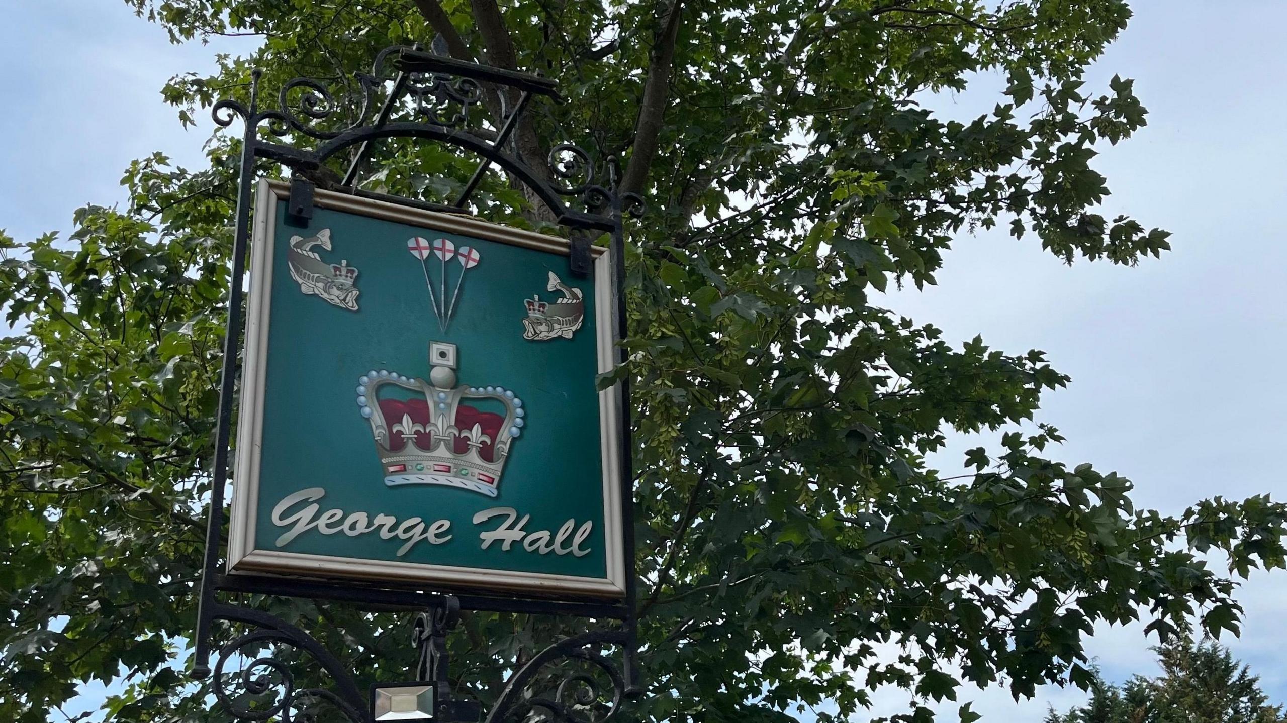 A green sign is held up by a tall black frame in front of a tree. The sign says "George Hall" and features a crown, fish and three darts.