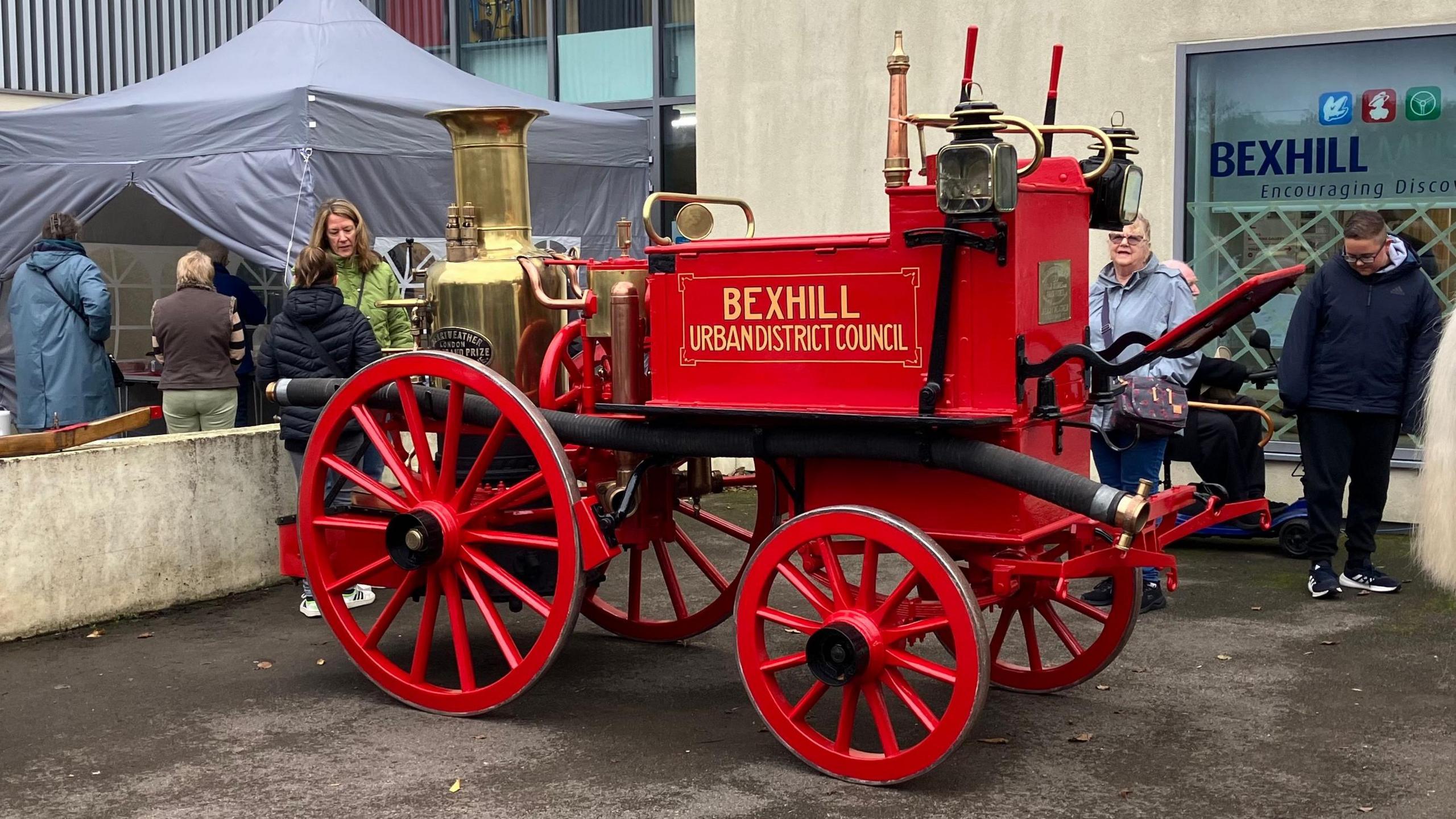 A red cart-like vehicle, with a big red wheel at the front and a smaller red one at the back. There is a boiler and other pieces of machinery on top of the cart 